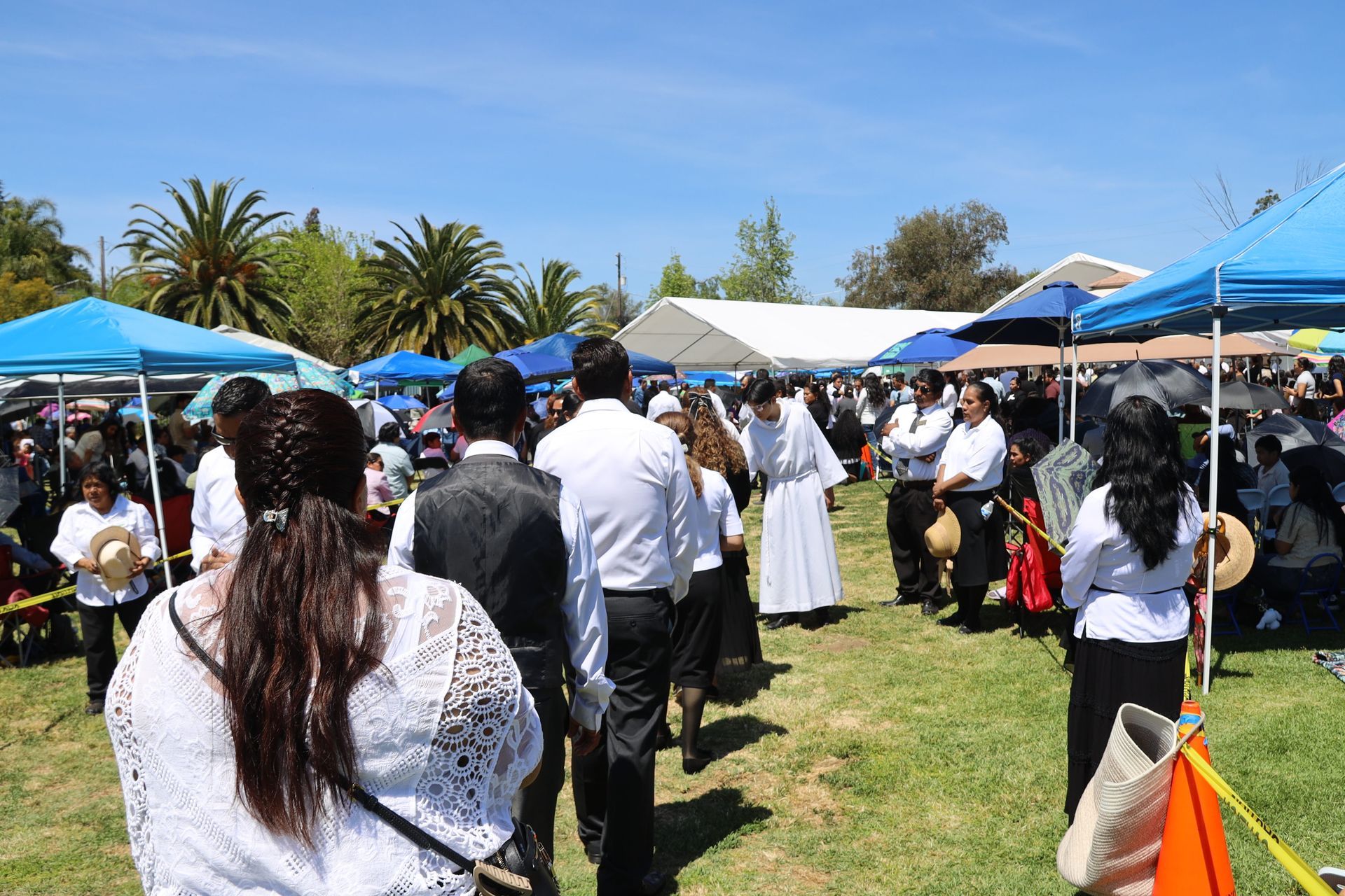 Outdoor gathering with people in white attire under blue tents on a sunny grassy lawn.