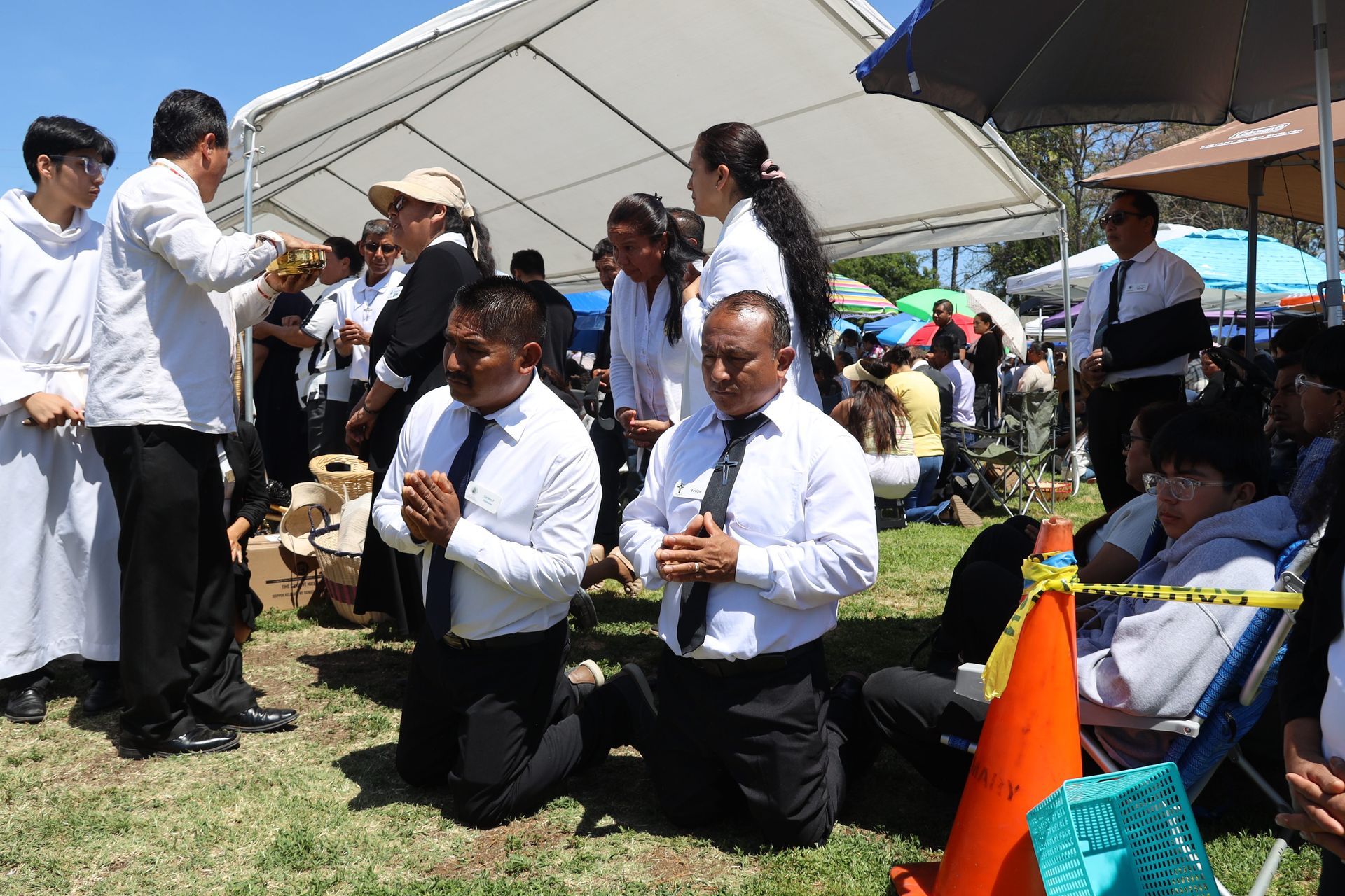 People kneel and play instruments at an outdoor event under a white tent.