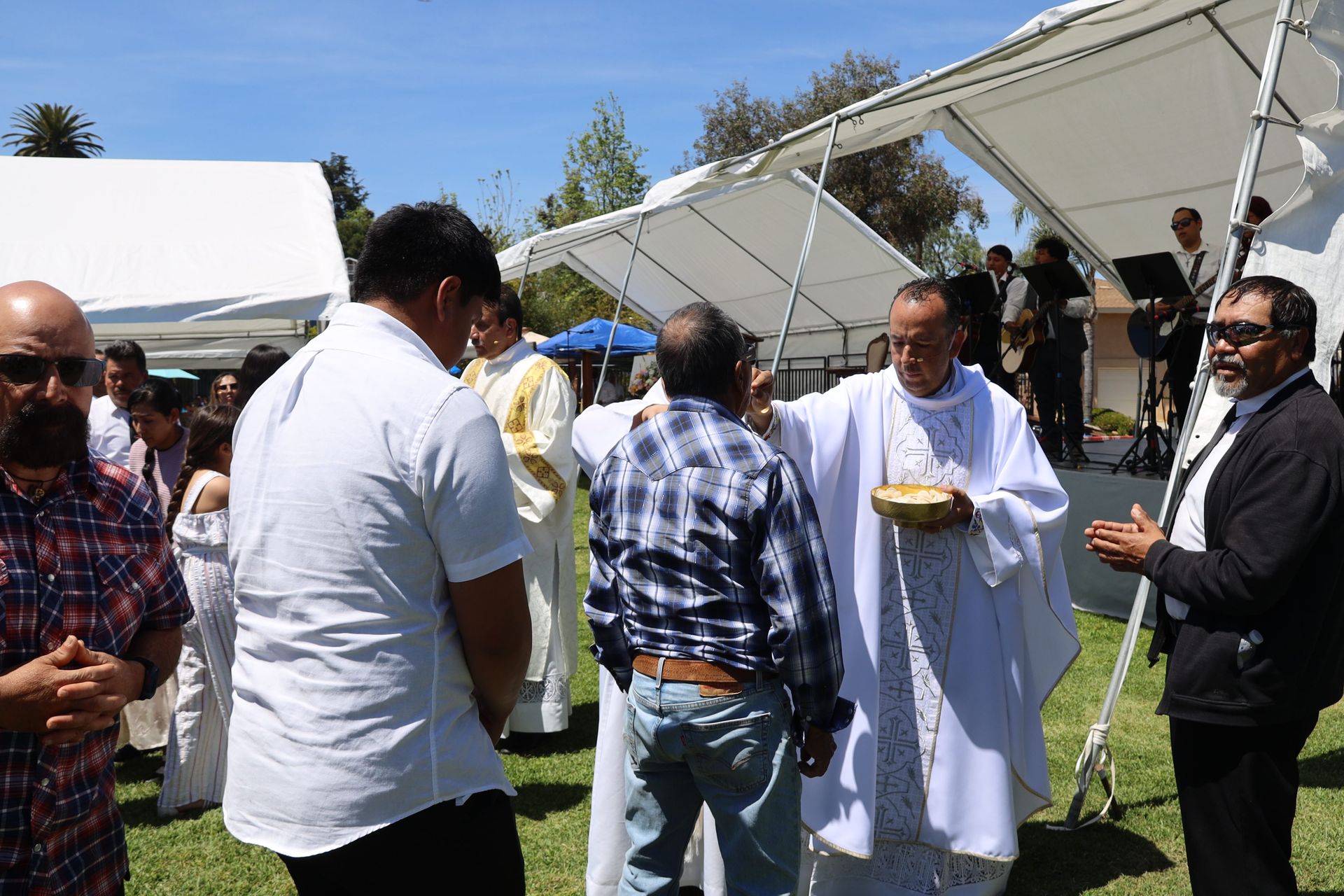 People gathered outdoors near a white canopy tent, one man holding a bowl and another speaking in white robes.
