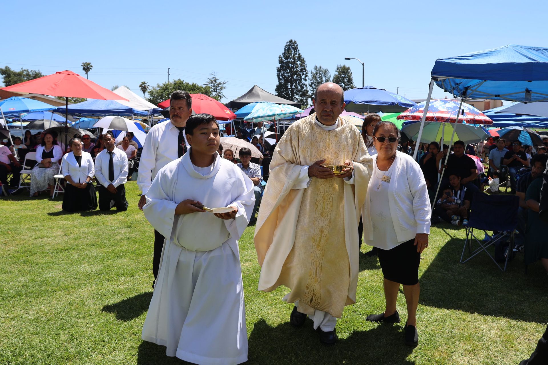 Clergy in white robes posing outdoors at a crowded event with colorful tents and umbrellas behind them