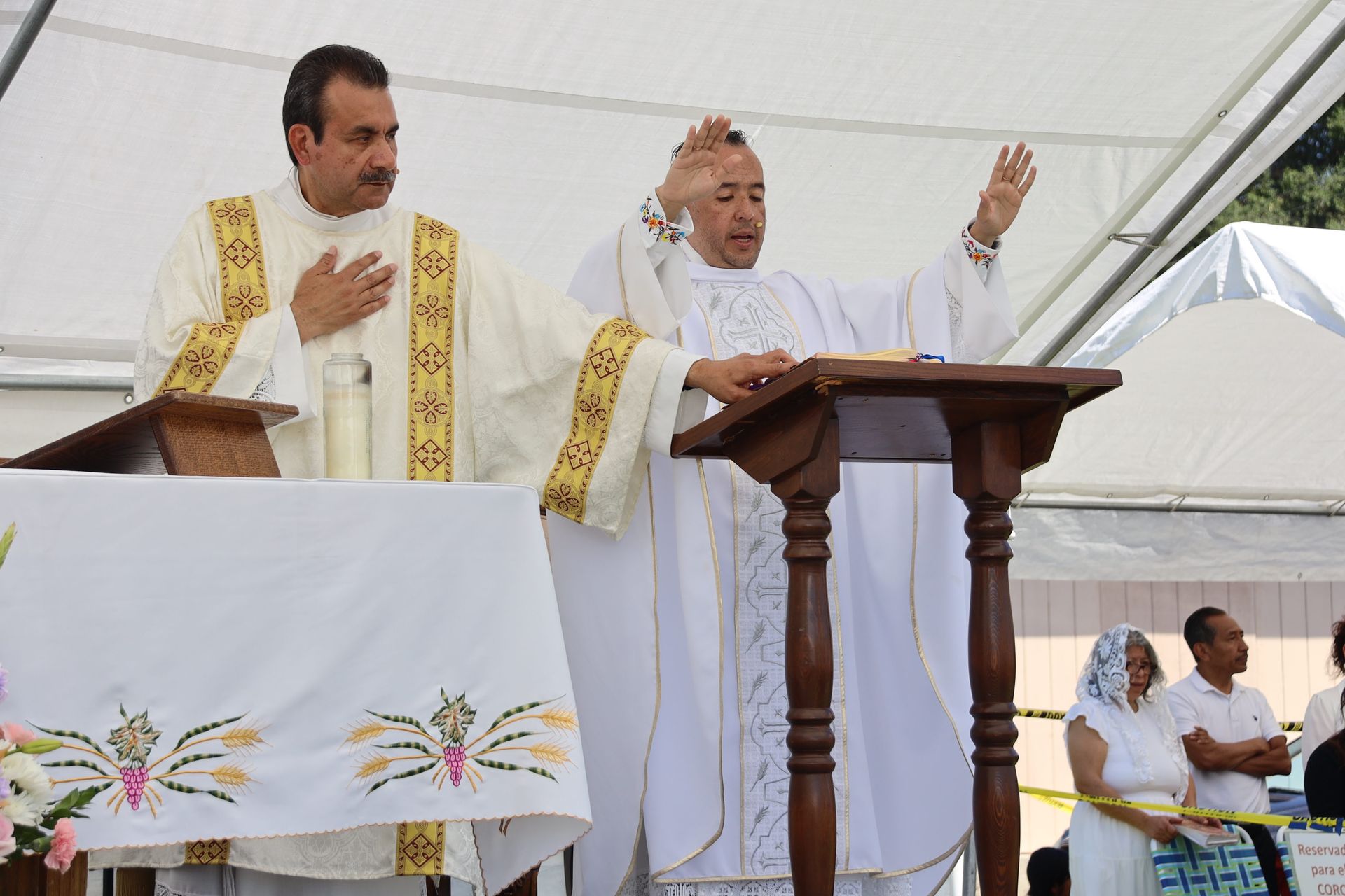Two priests in white and gold vestments speak at a church altar under a tent.
