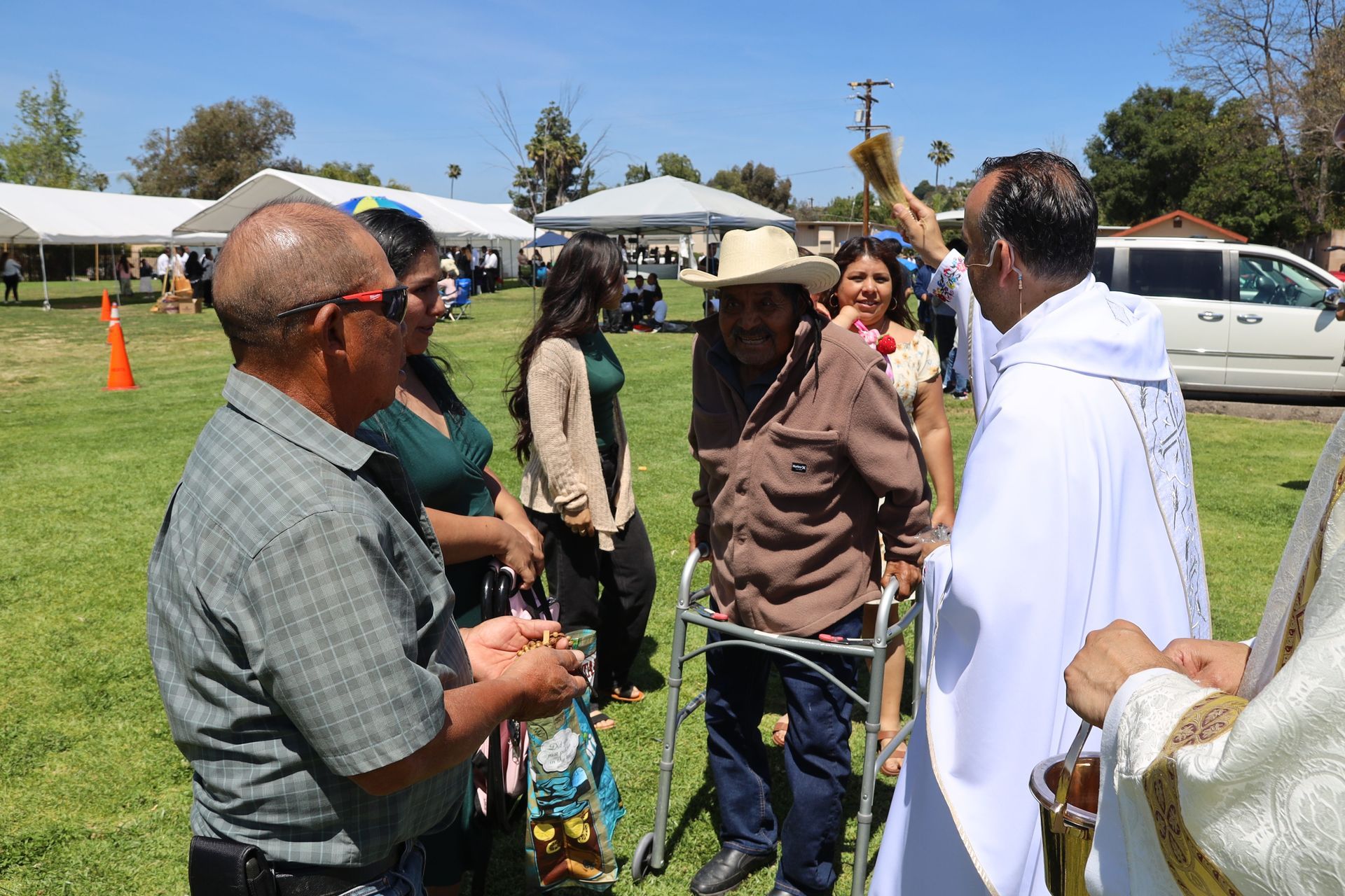 People chatting outdoors on a sunny lawn near parked cars and white canopies