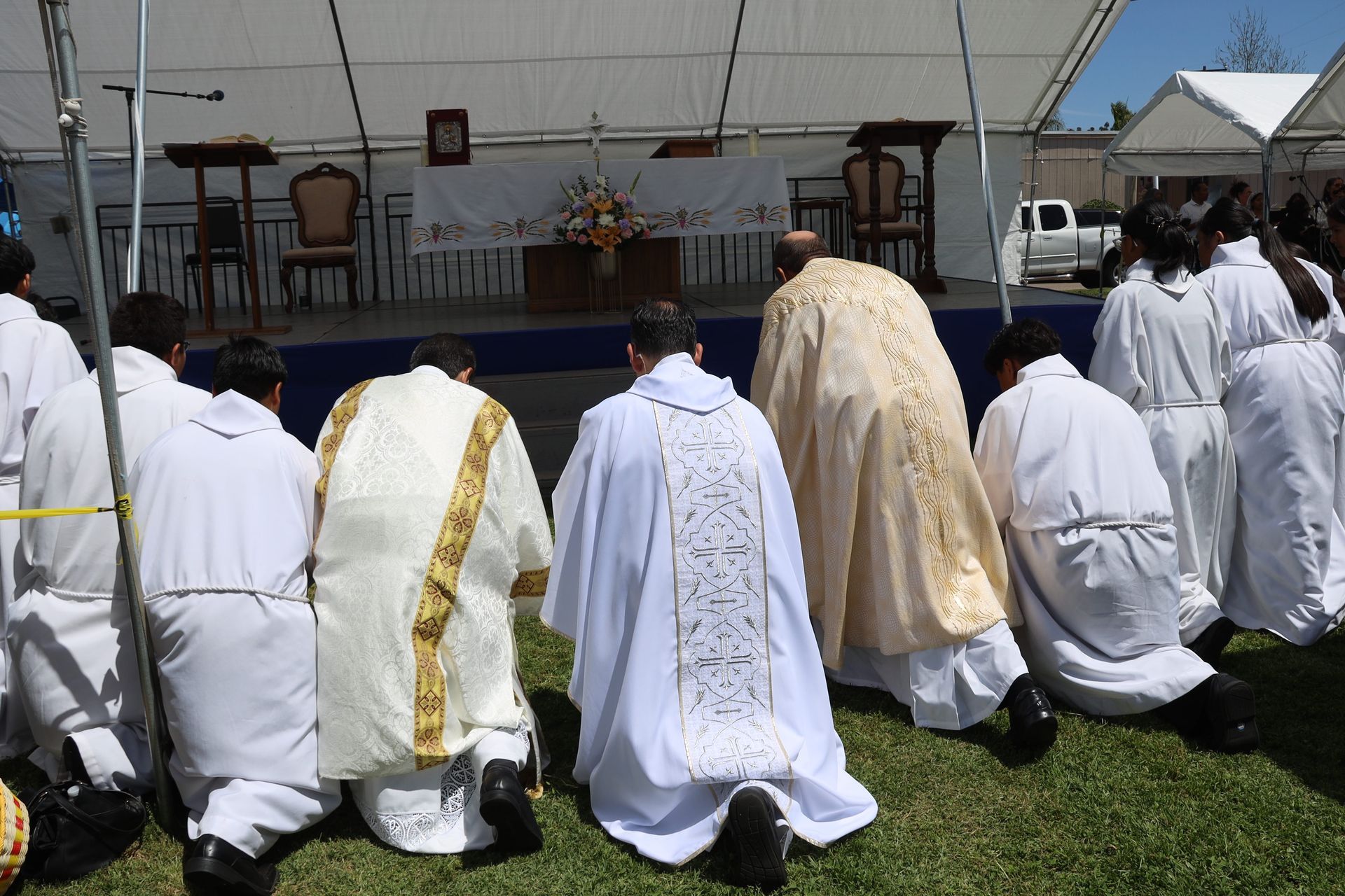 People in white robes kneeling on grass during an outdoor ceremony under a tent