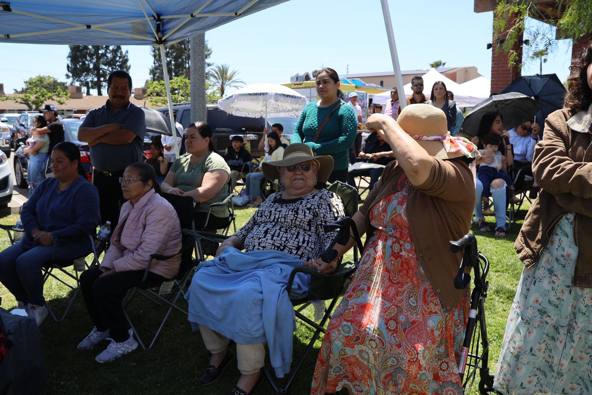 People seated under a canopy at an outdoor gathering, with one person shading their eyes in the sun.