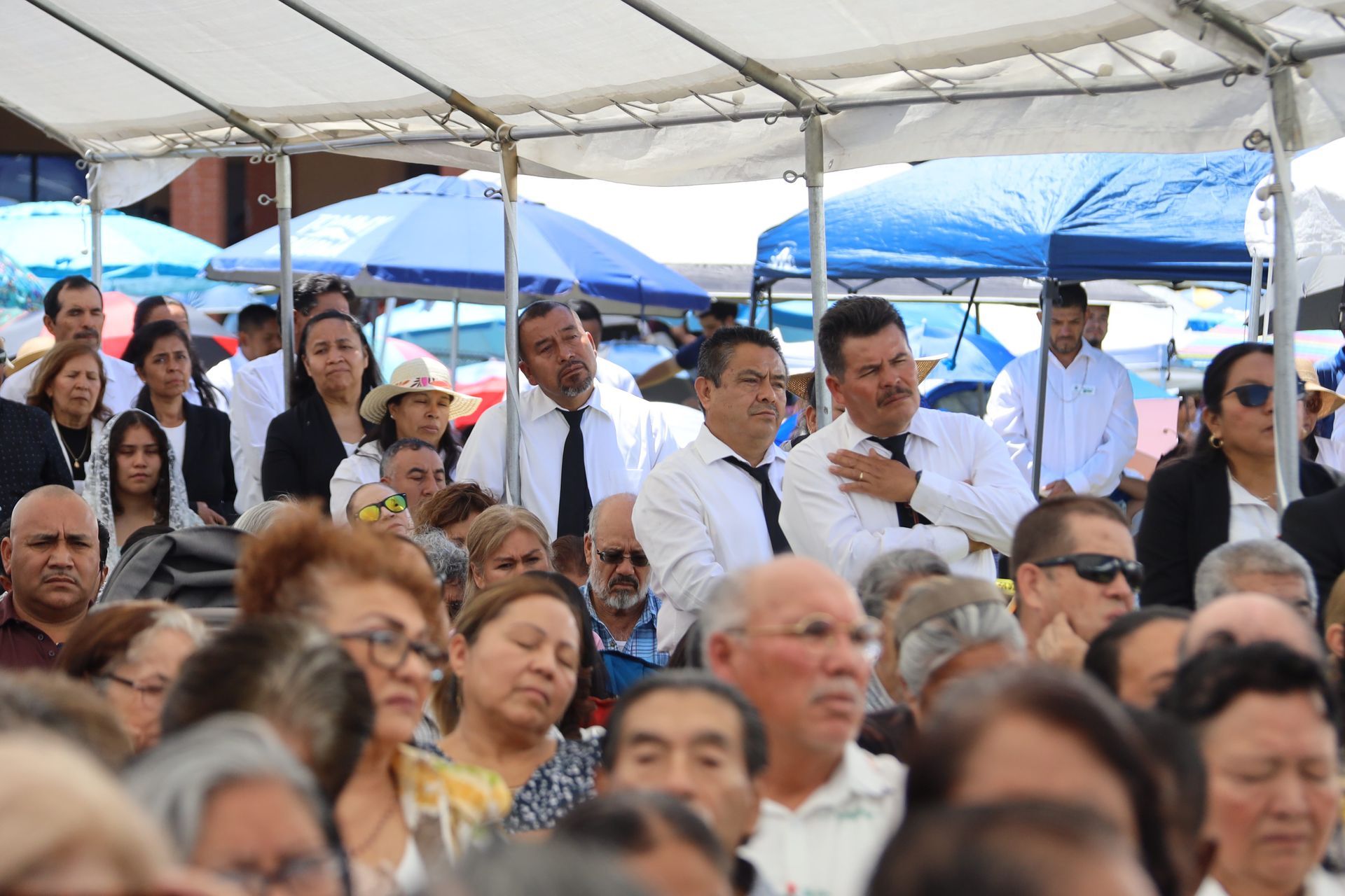 Crowd seated under a canopy, with several people in white shirts and blue umbrellas in the background
