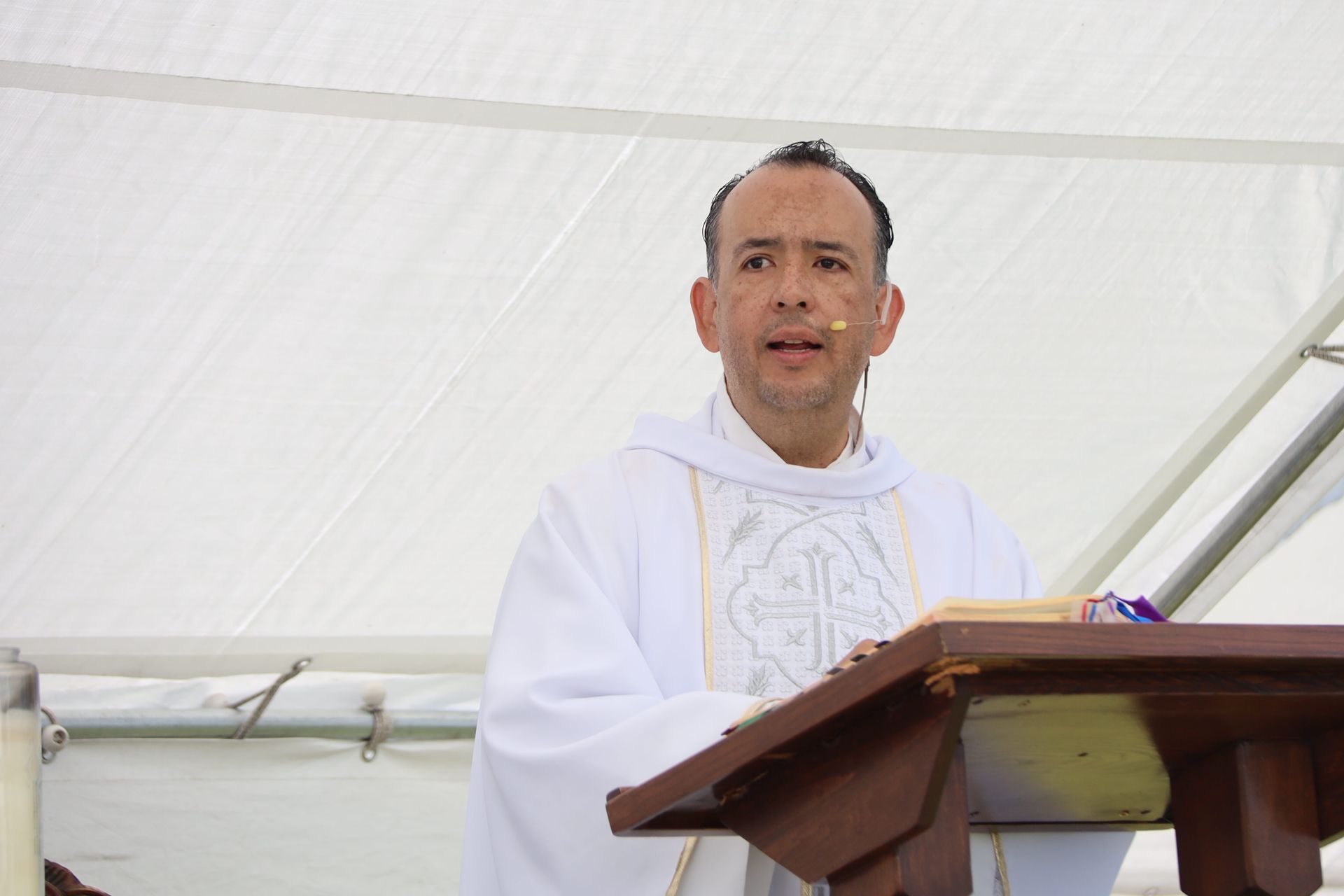 Person in white vestments speaking at a wooden lectern under a white tent.