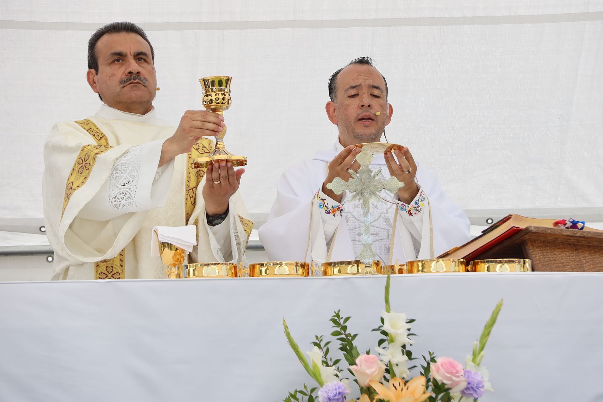 Two priests in white robes celebrate Mass at an altar, one holding a chalice and the other prayerfully clasping hands.