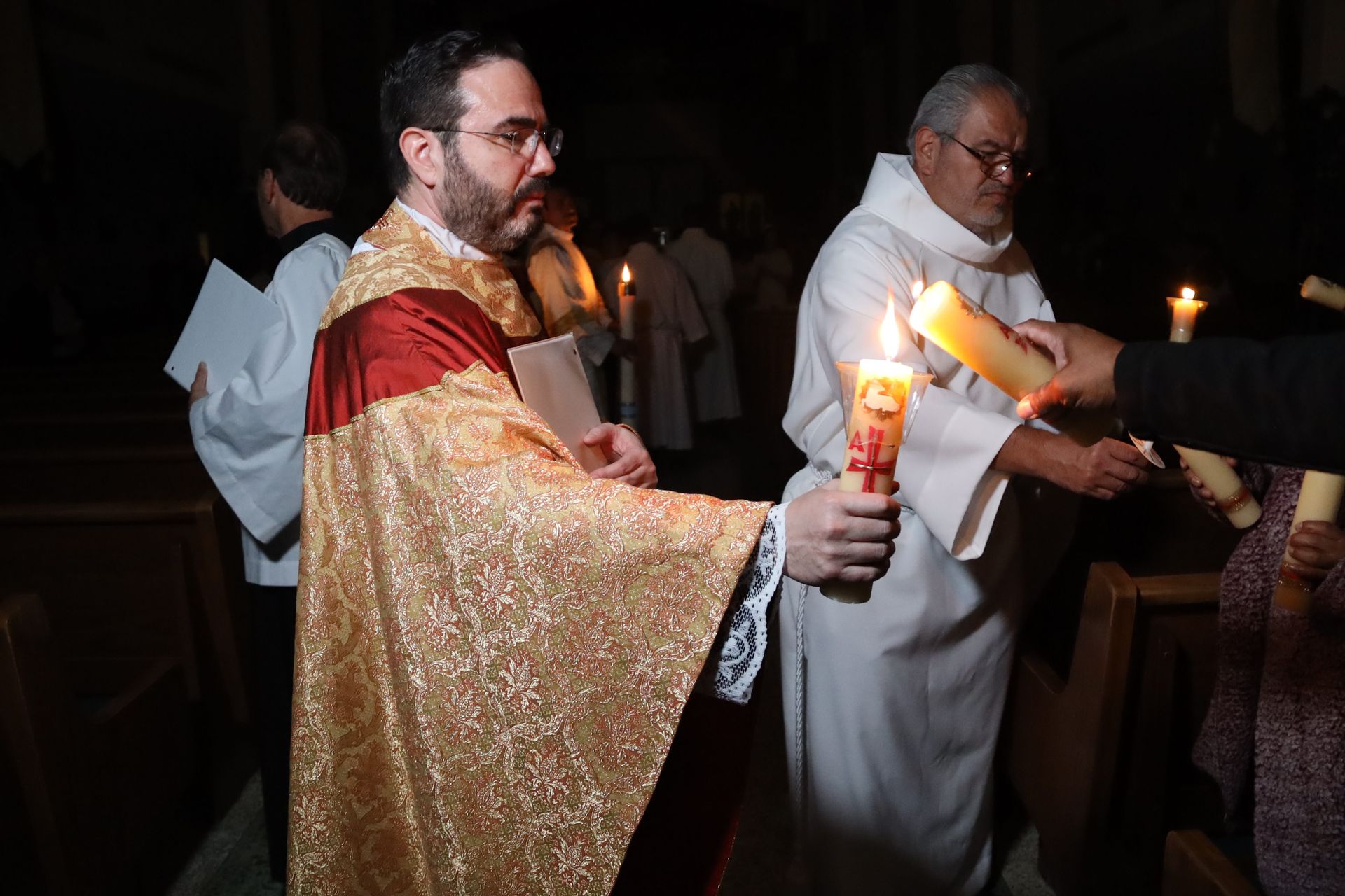 Clergy in white and gold robes holding lit candles during a dim church procession