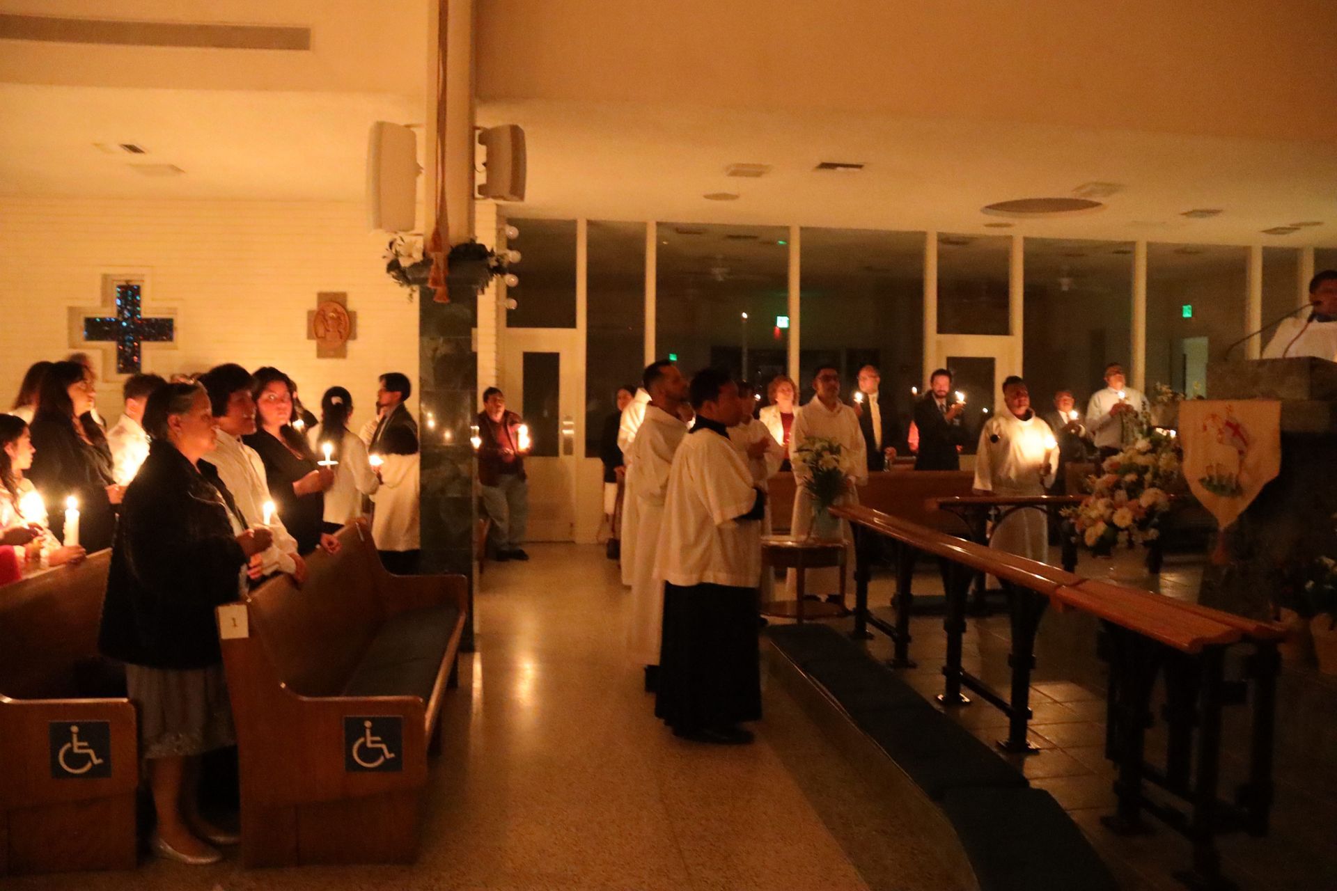 Candlelit church service with people standing in pews, holding candles in a dim chapel.