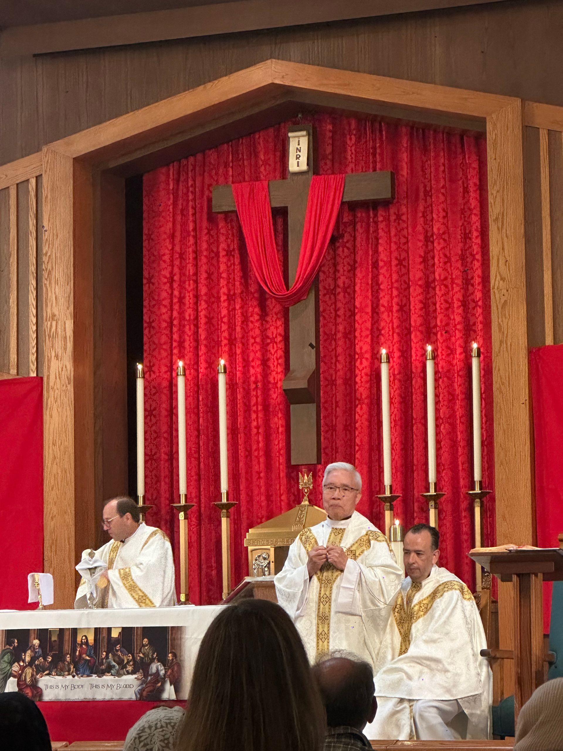 Clergy in white robes seated at an altar with red drapery and a large cross in the background