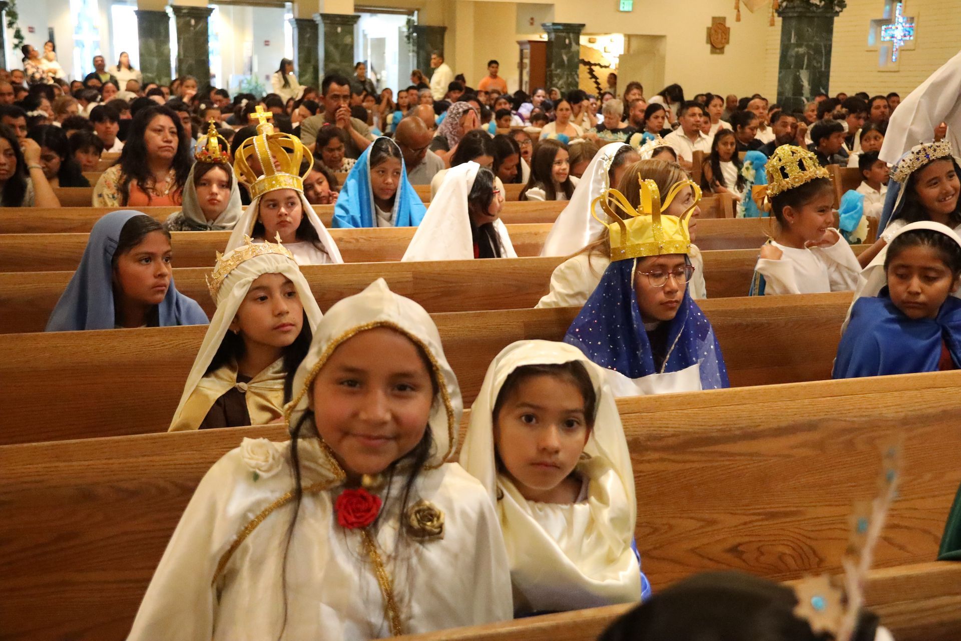 Children in white robes and gold crowns seated in church pews during a religious ceremony