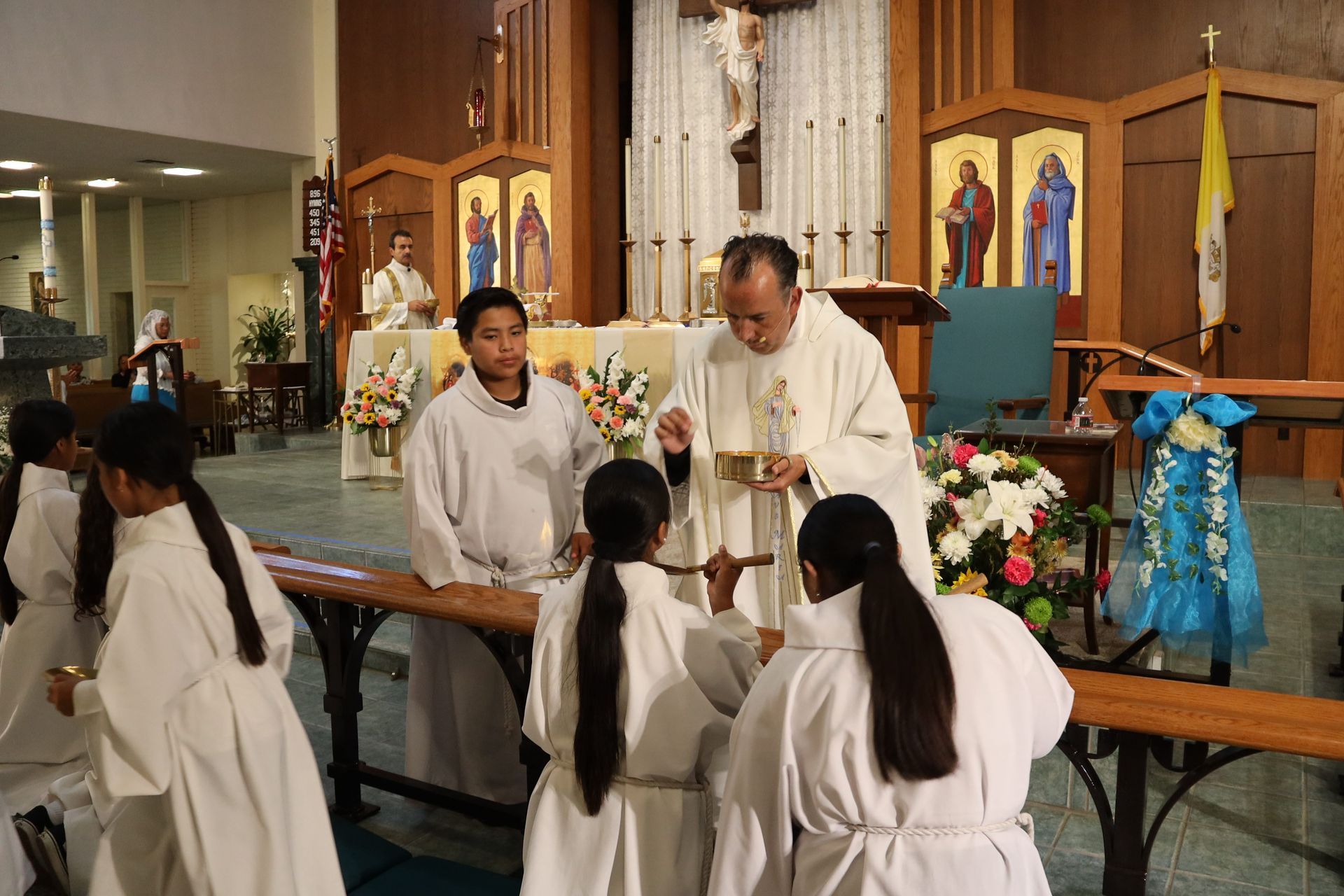 Priest blessing kneeling children at church altar, with white and blue robes and floral decorations