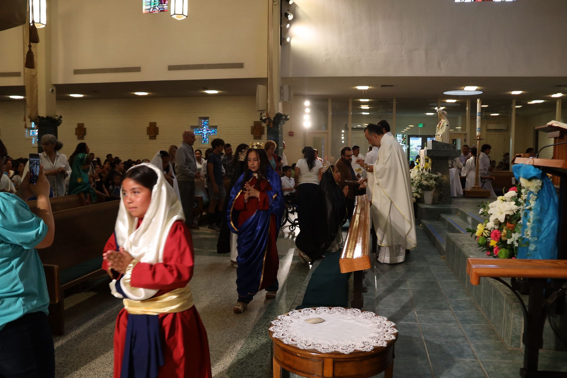People in colorful robes gathered indoors for a church ceremony, with a decorated table in the foreground.