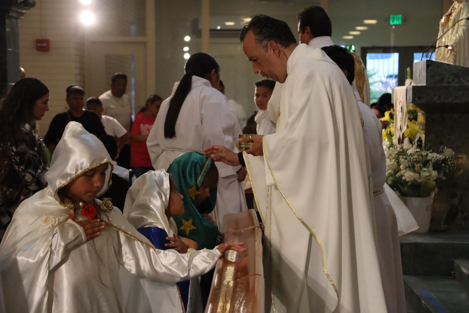 People in white robes and festive clothing gather indoors for a religious ceremony.