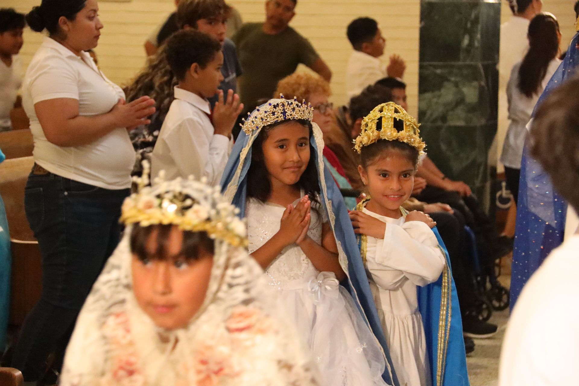 Children in white costumes with gold crowns at a festive indoor ceremony, some with hands clasped in prayer