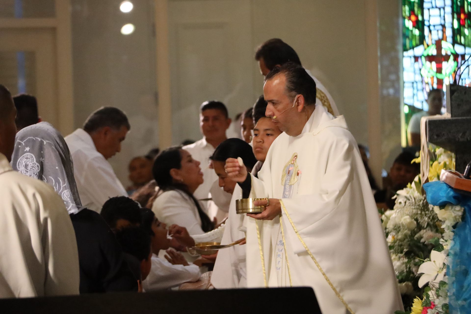Priest in white vestments holds a chalice during a church service, with congregants behind him