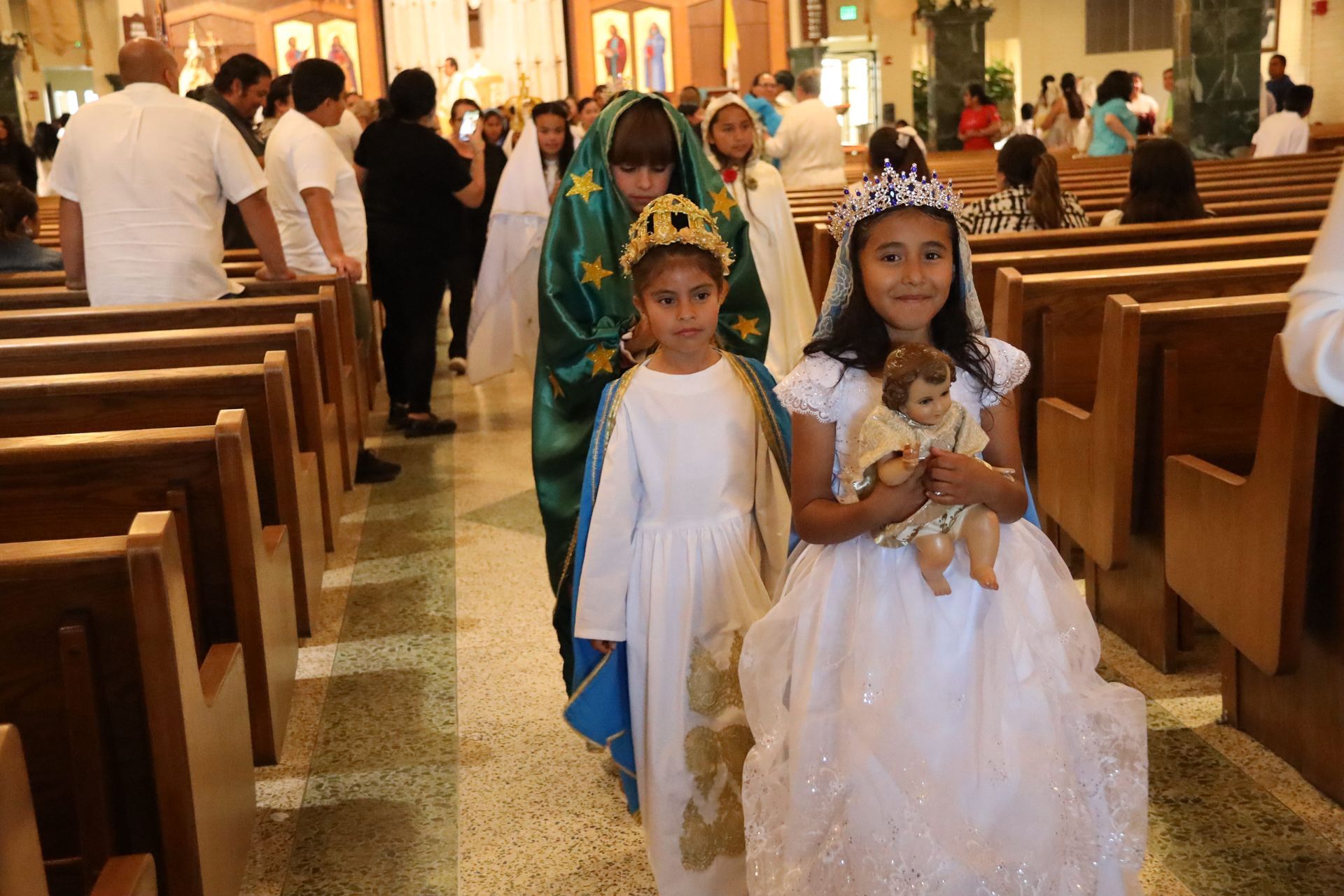 Children in white and blue ceremonial outfits walking down a church aisle, one holding a doll.