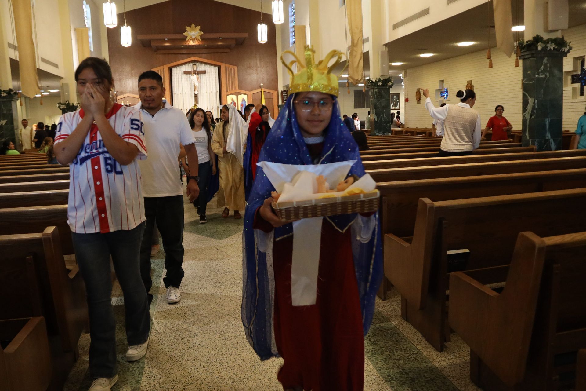 Two children in costumes walk down a church aisle during a procession, one carrying a tray.