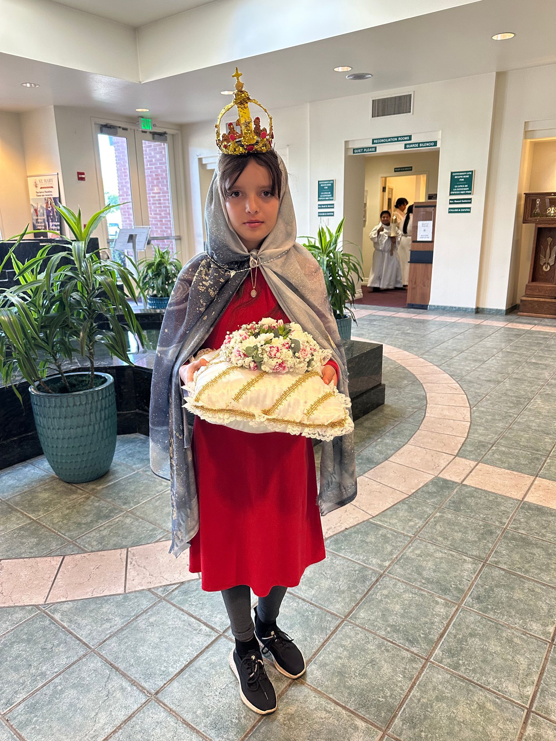 Child in a red dress and gray cape holding a decorated bread tray in a bright lobby