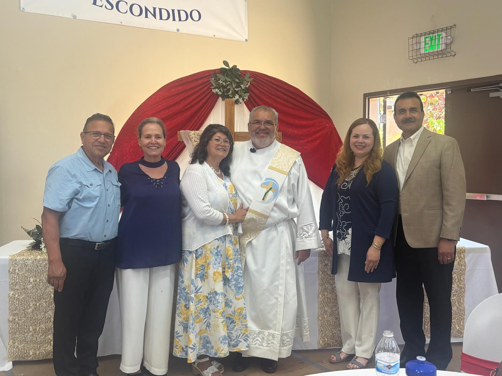 Group posed on a stage in front of a red draped arch, with a central officiant in white robes.