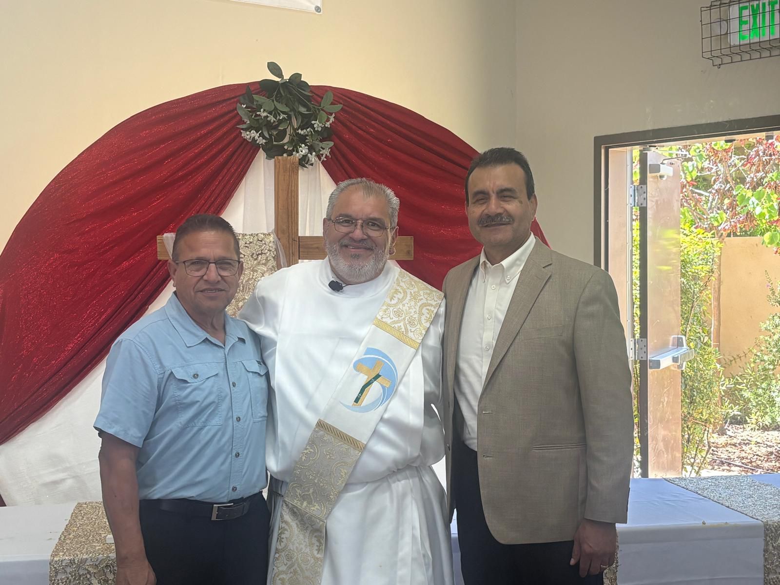 Three men standing indoors at a decorated event, smiling in front of red drapery.