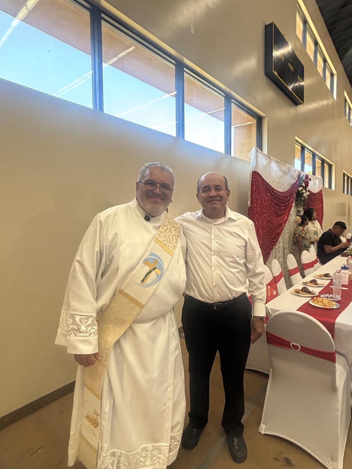 Two men standing together at a decorated indoor event table, one in white robes and gold sash, smiling.
