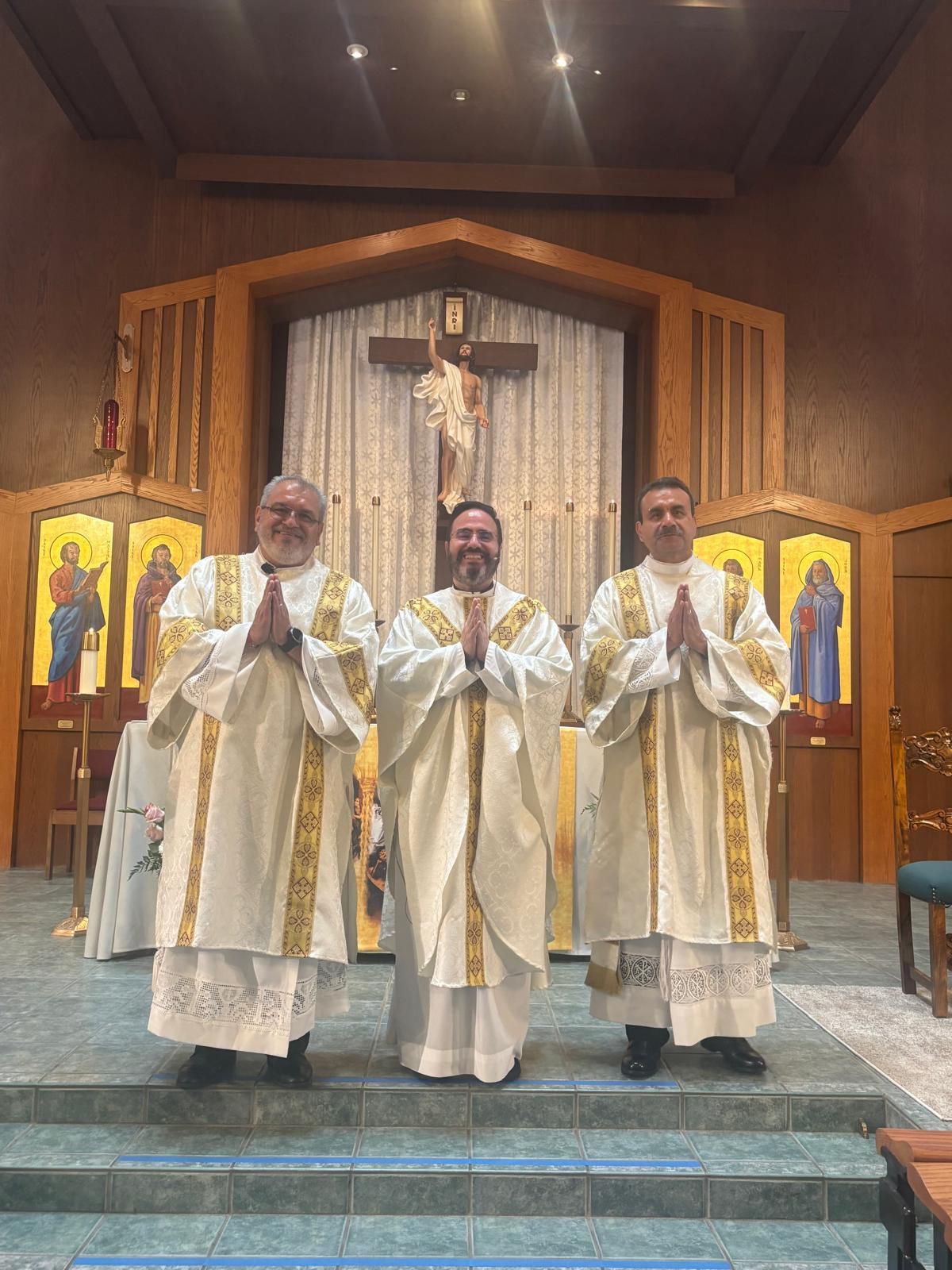 Three clergy in white robes stand before an altar, with a crucifix and icons in the church background.