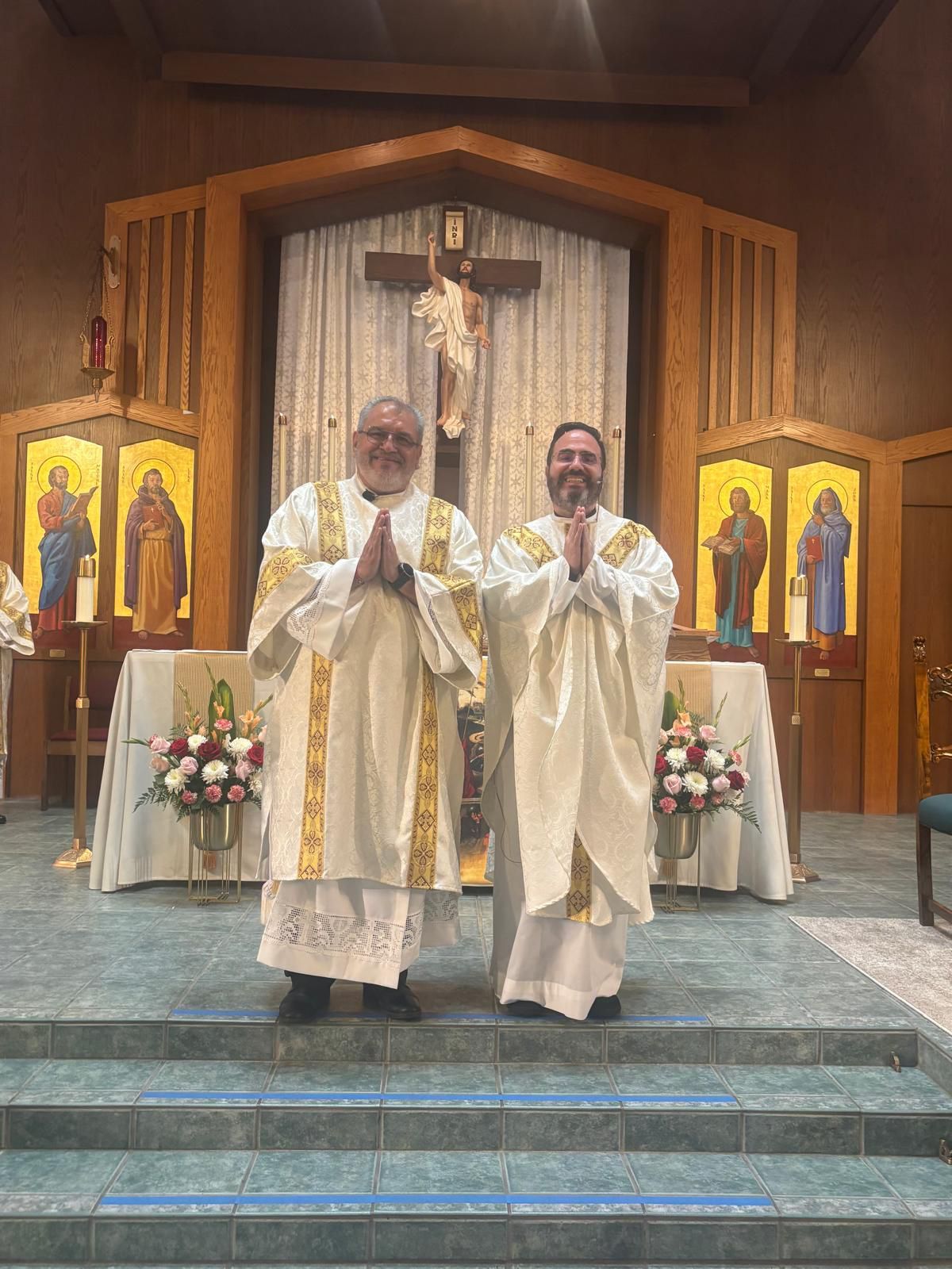 Two priests in white vestments stand at the altar in a church, with a crucifix and icons behind them