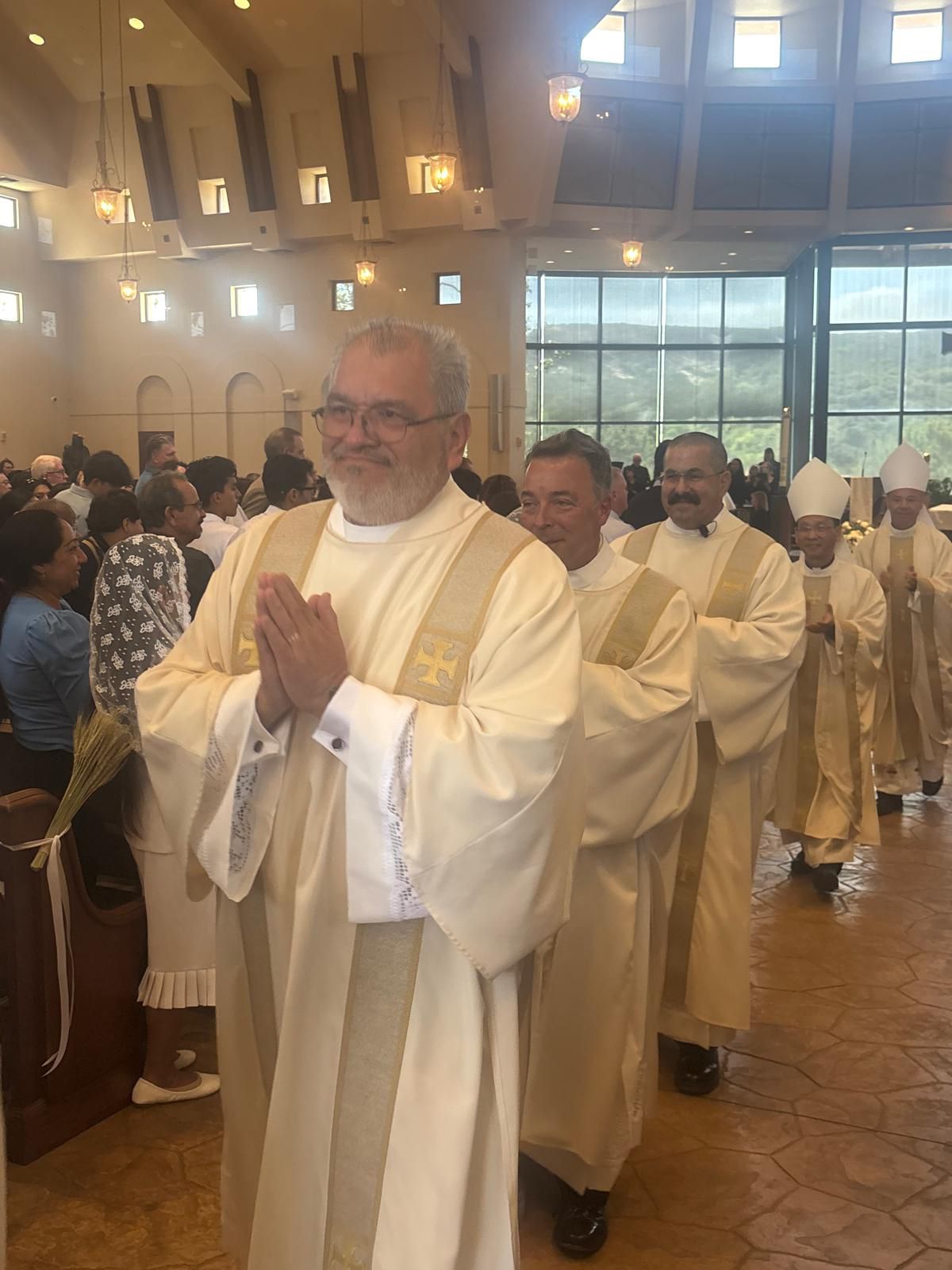 Clergy in gold vestments process through a church aisle, with the lead priest smiling and hands clasped.