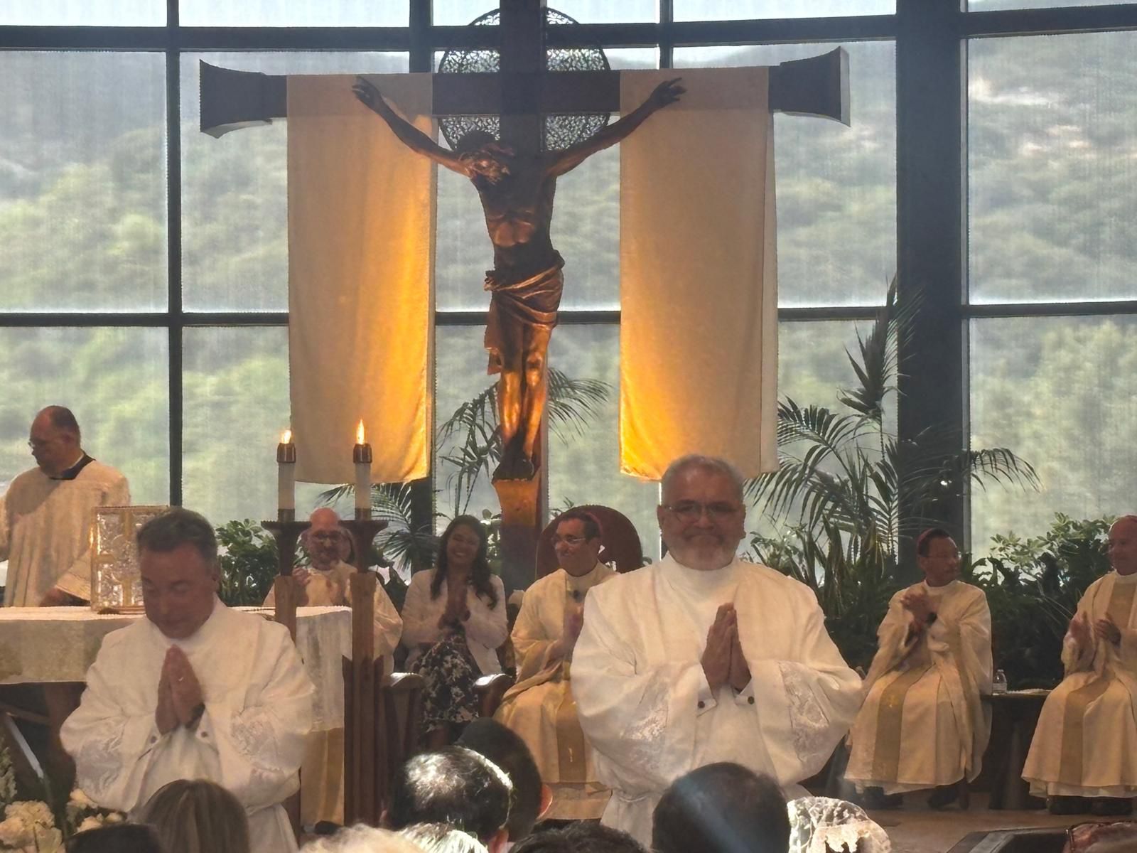 Church altar with crucifix, candles, and seated clergy before large windows