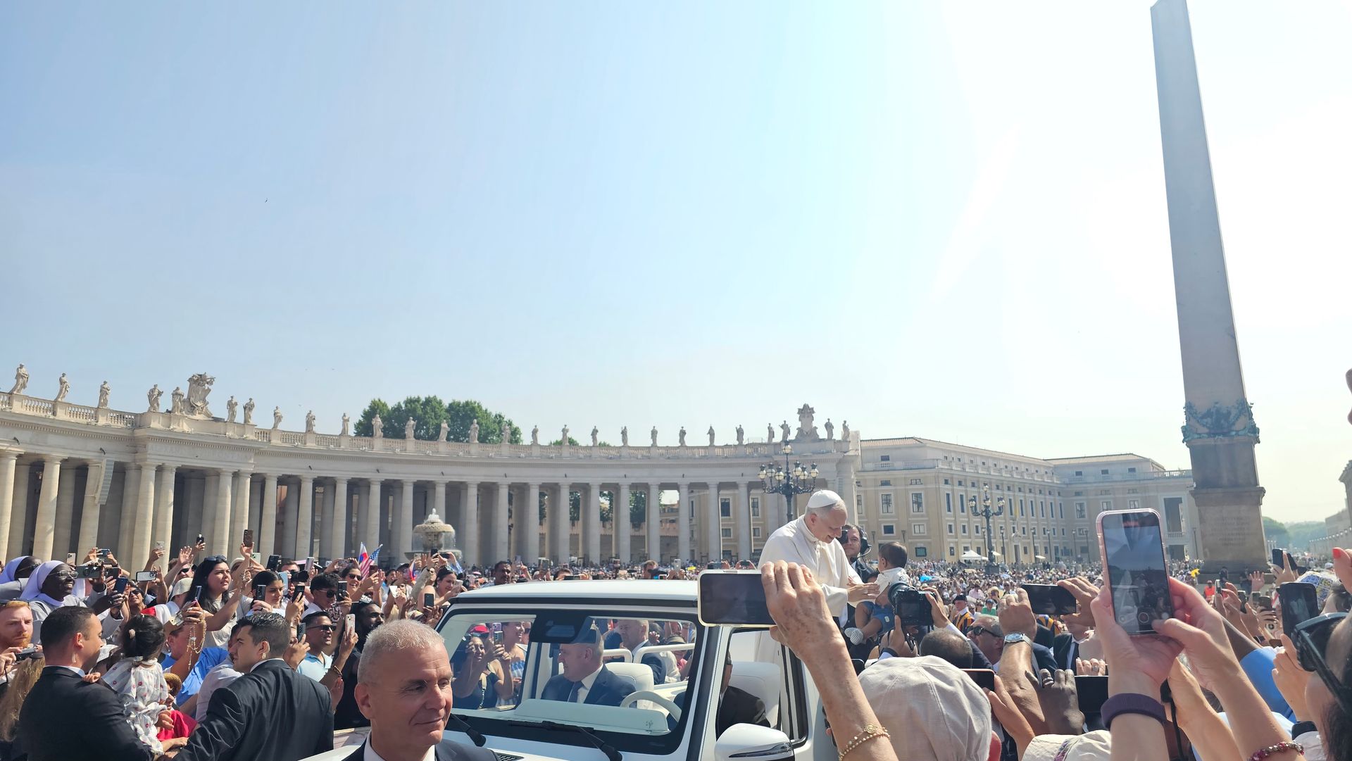 Crowd in St. Peter’s Square with popemobile and Vatican colonnade under bright blue sky