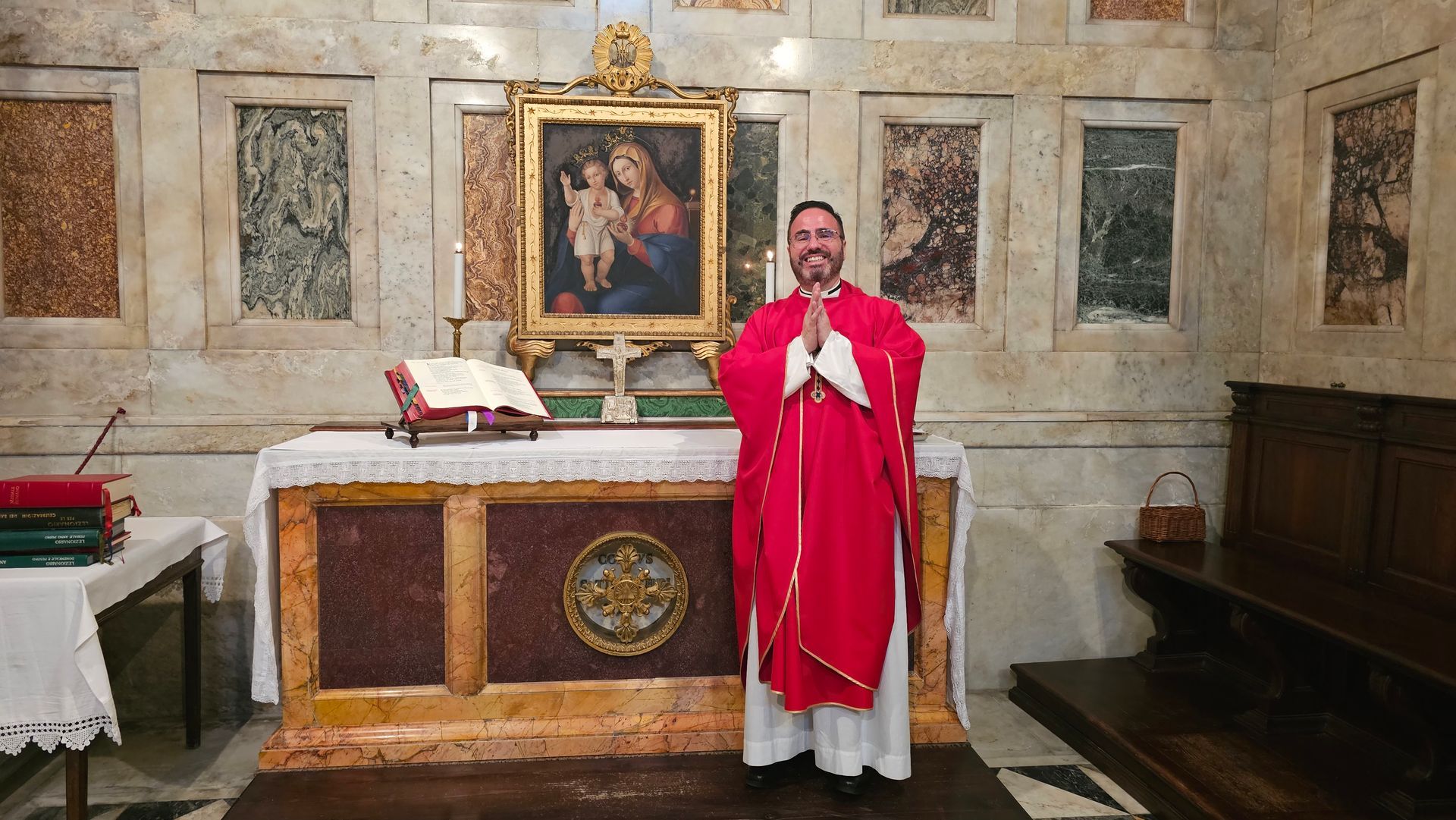 Priest in red vestments standing before a church altar with an open book and a portrait behind him