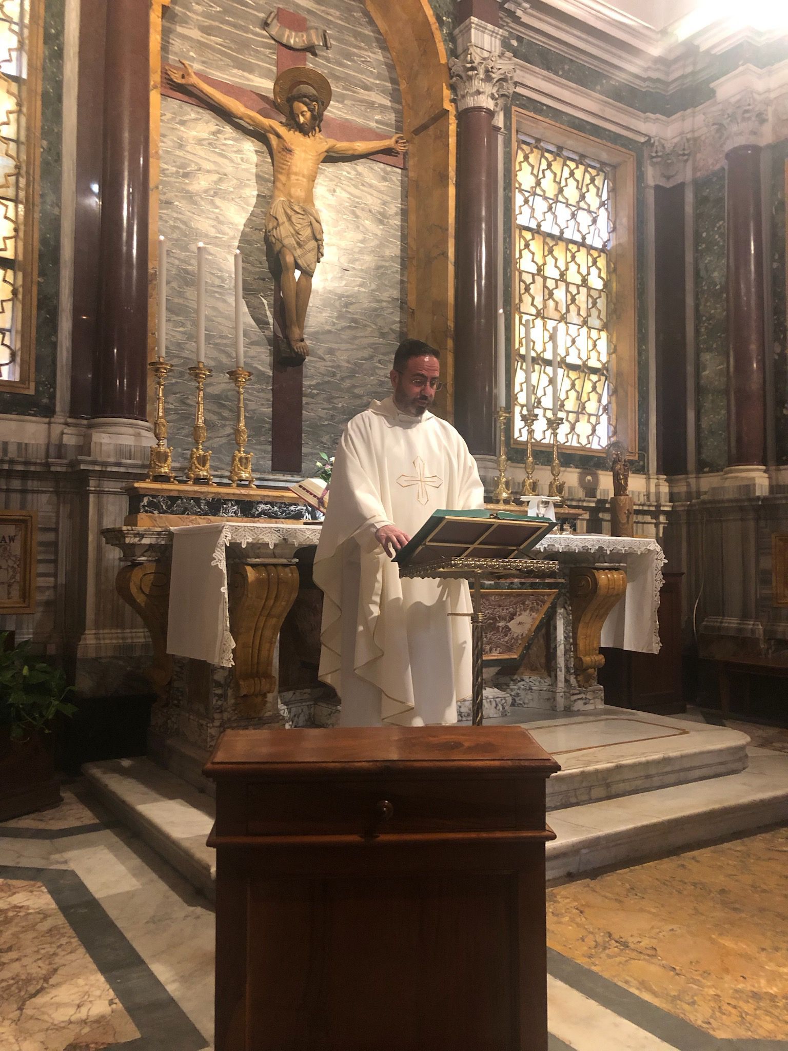 Person in white robe speaking at a lectern inside an ornate church with a crucifix behind.
