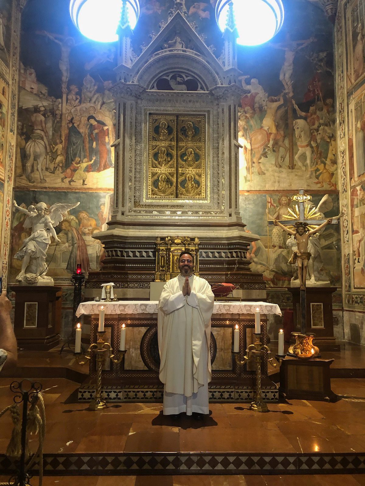 Priest in white vestments standing at an ornate altar inside a richly decorated church