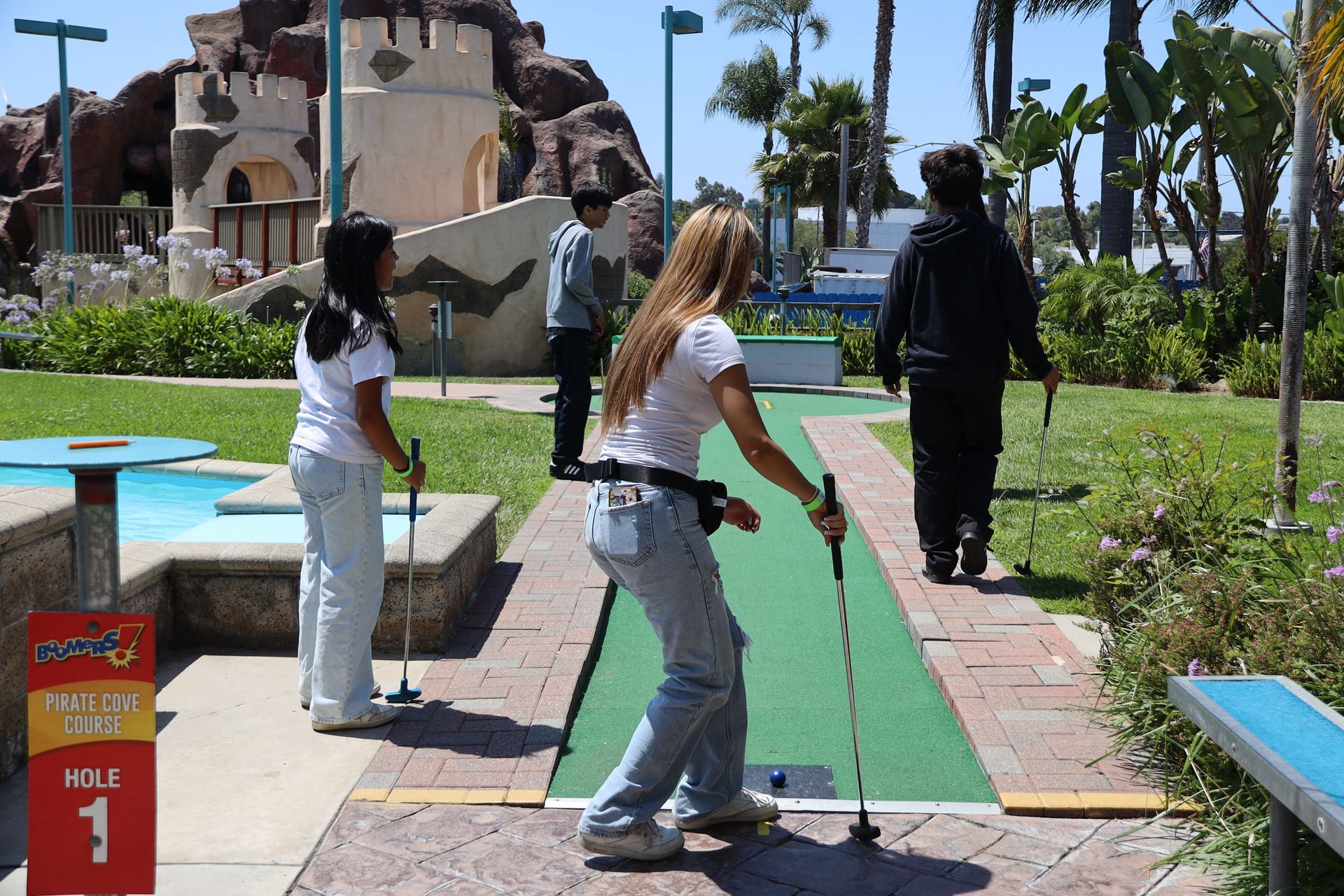 People playing mini golf on a green lane outdoors, with a castle-like building in the background.