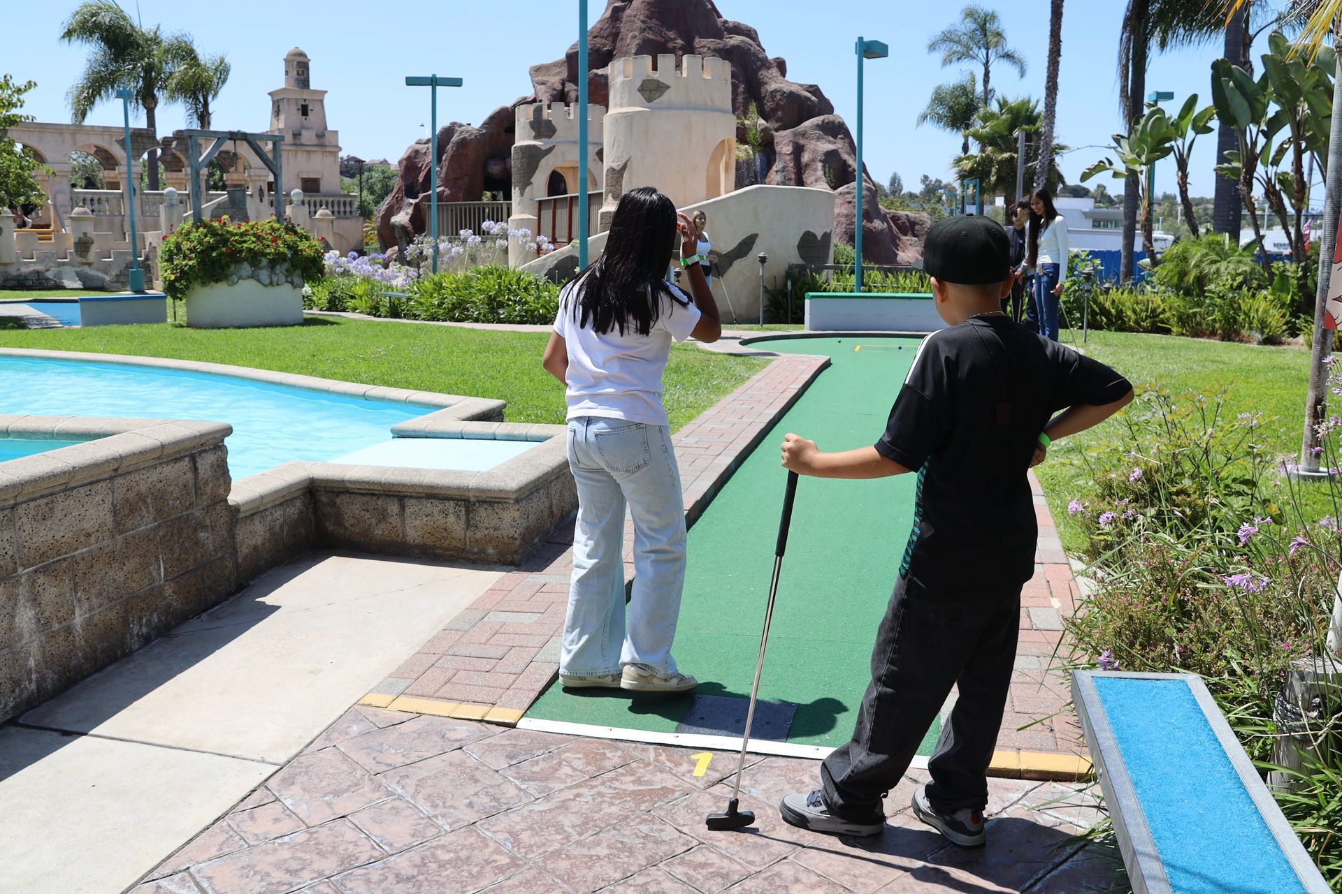 Two people playing mini golf beside a turquoise pool and resort buildings under palm trees
