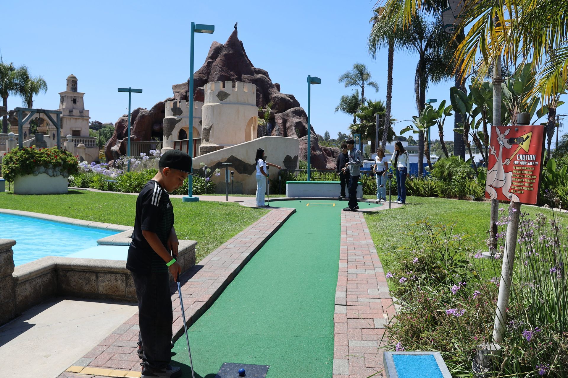 Mini-golf course with a green putting path, a worker in black, and a castle-themed structure in the background