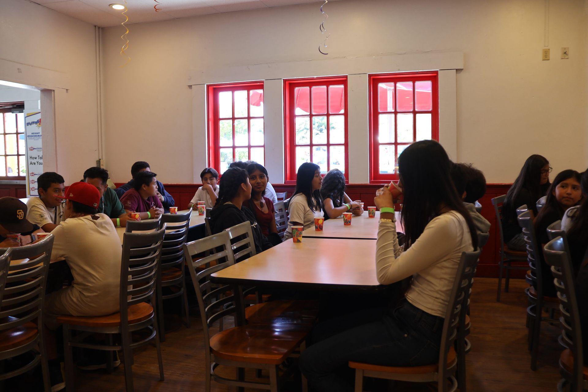 People seated around tables in a bright room with red windows, gathered for a meeting or meal