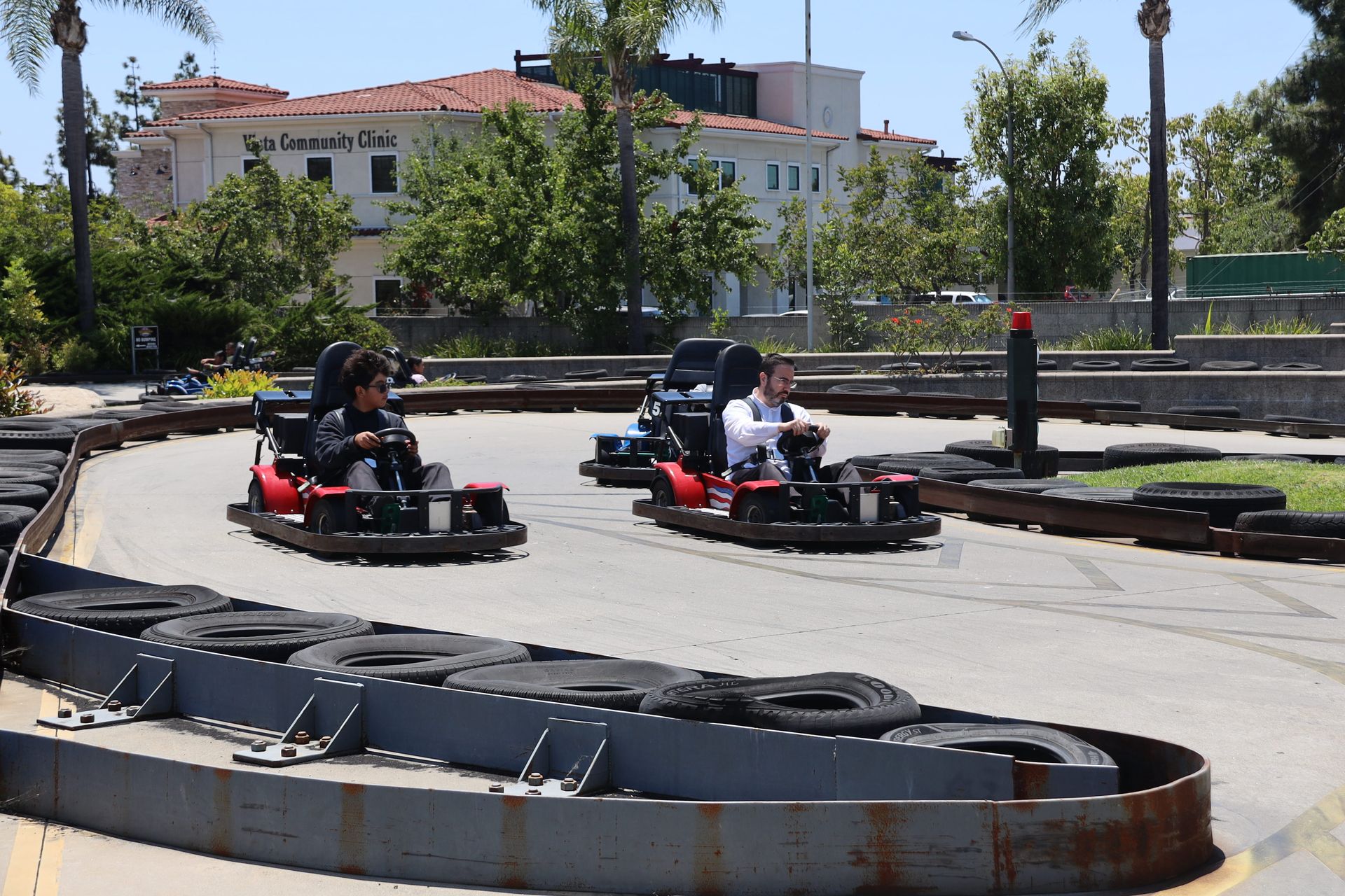 Two people driving go-karts around a sunny outdoor track with tire barriers