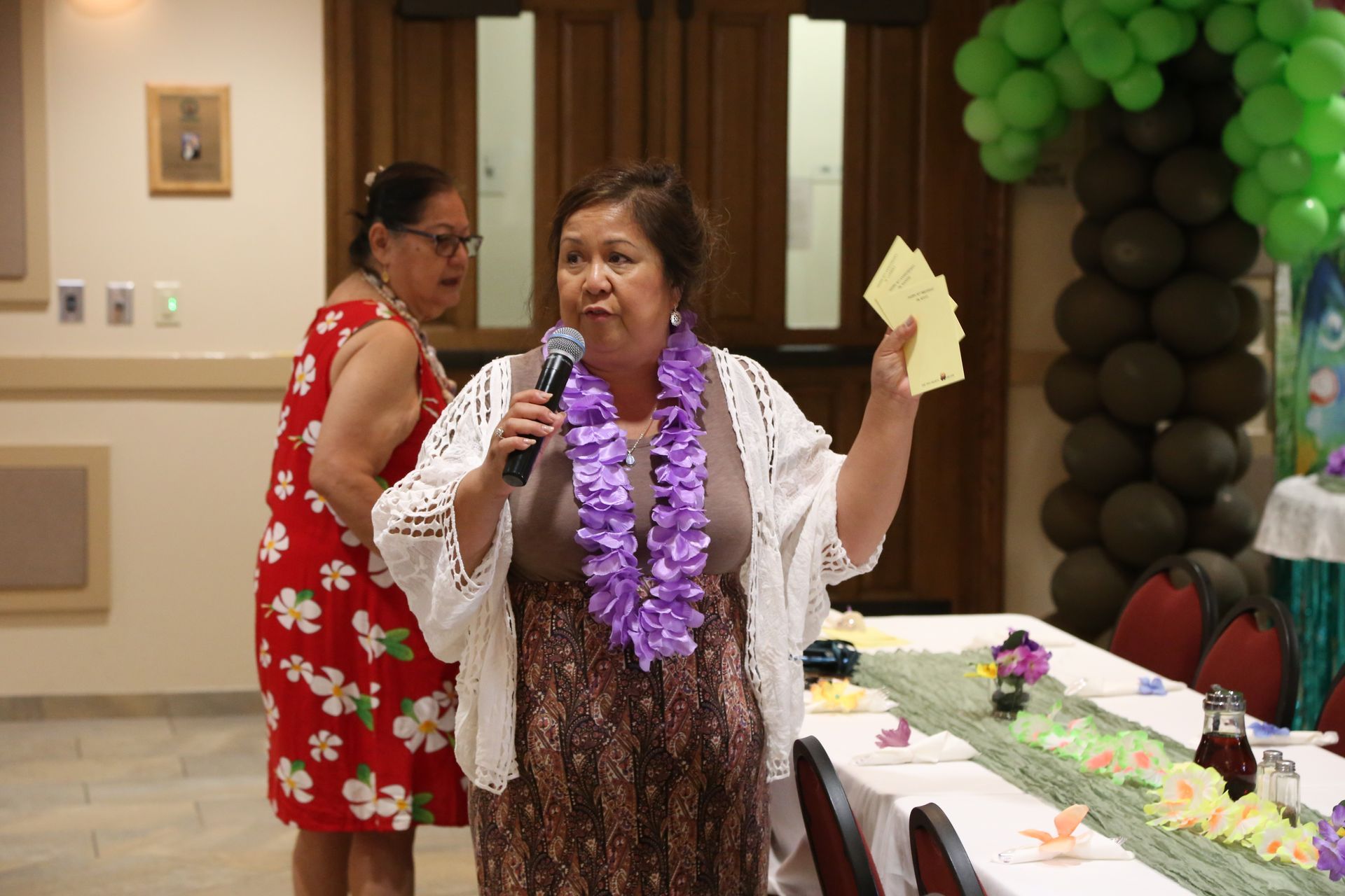 Woman speaking into a microphone at a decorated party table, holding cards, with green and brown balloons behind her