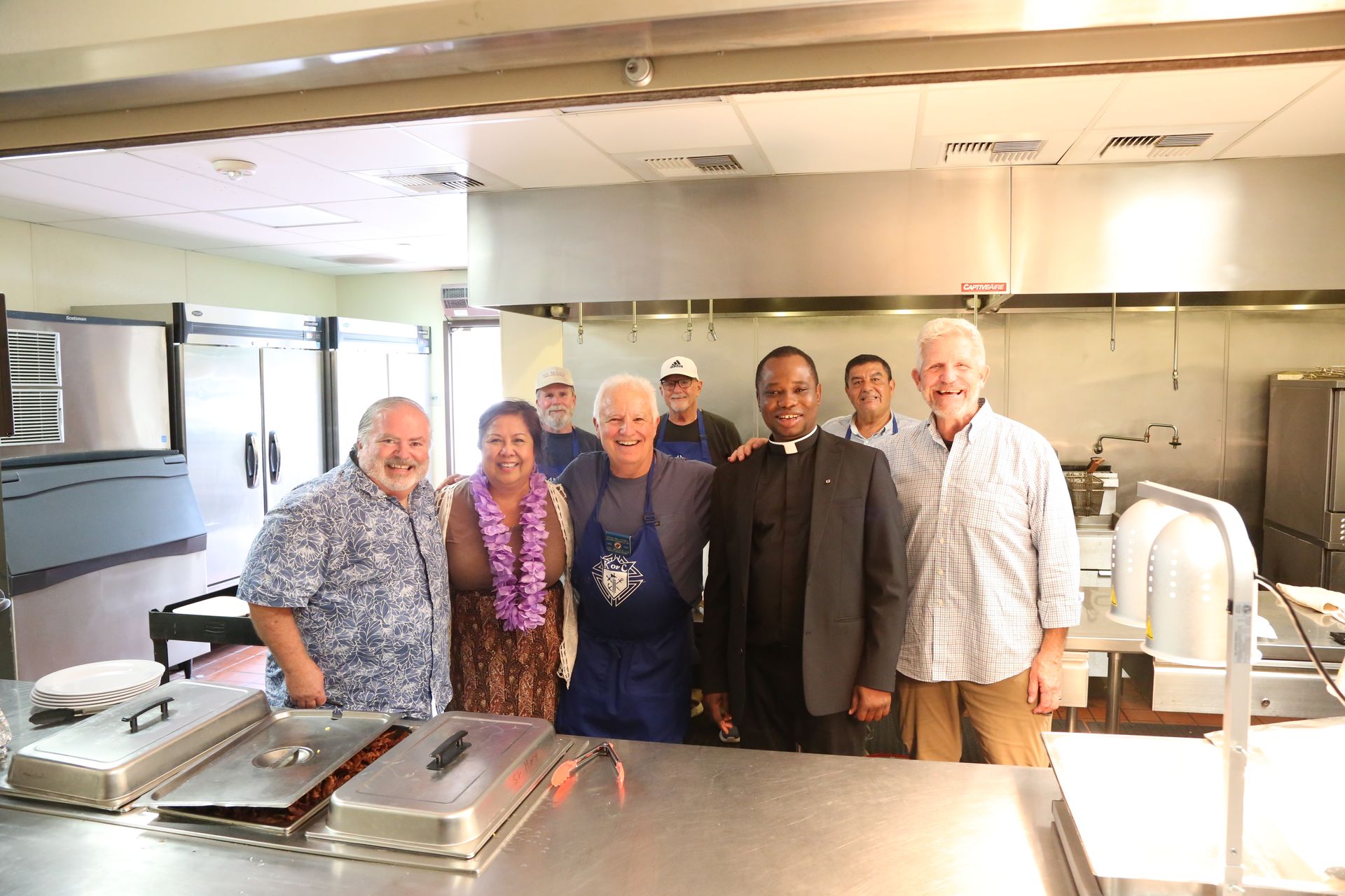 Group of six people standing in a bright commercial kitchen, smiling behind a stainless steel counter