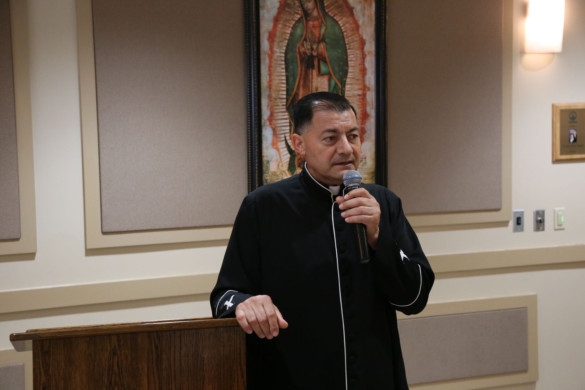 Man speaking into a microphone at a podium in a conference room, with a religious painting behind him