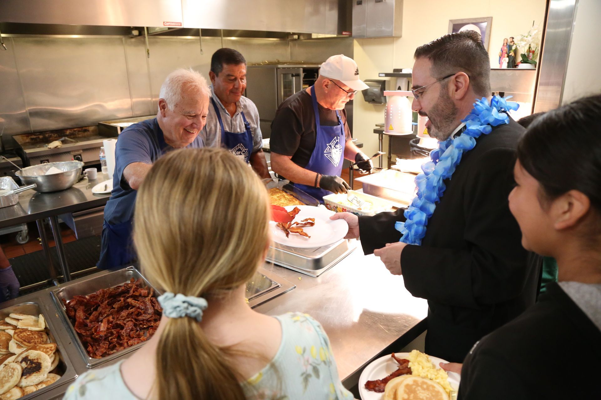 People in a kitchen serving food at a buffet counter, with trays of dishes and stainless steel equipment.