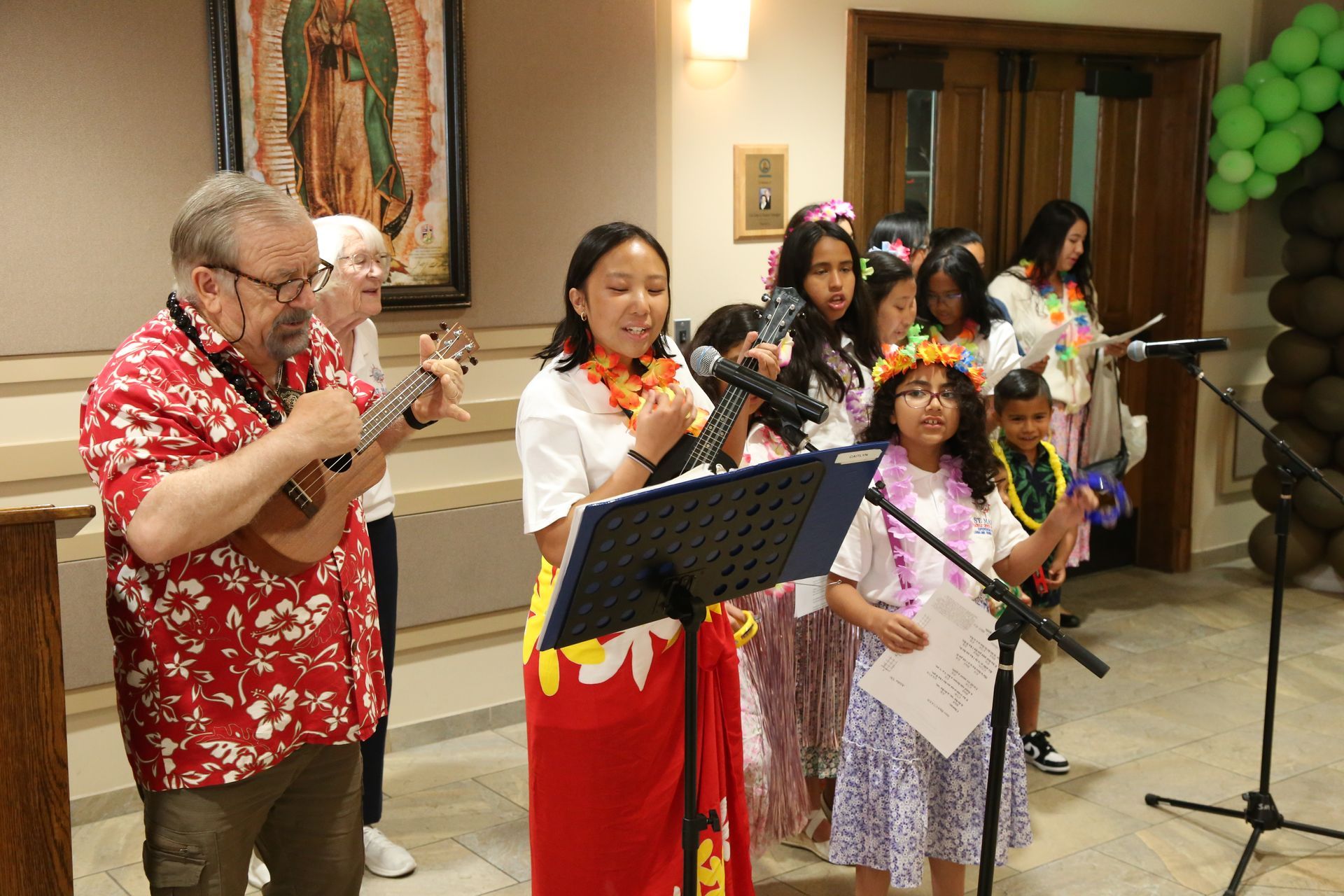 Group singing at microphones in a hall, with leis and colorful outfits, while a man in a red shirt claps nearby