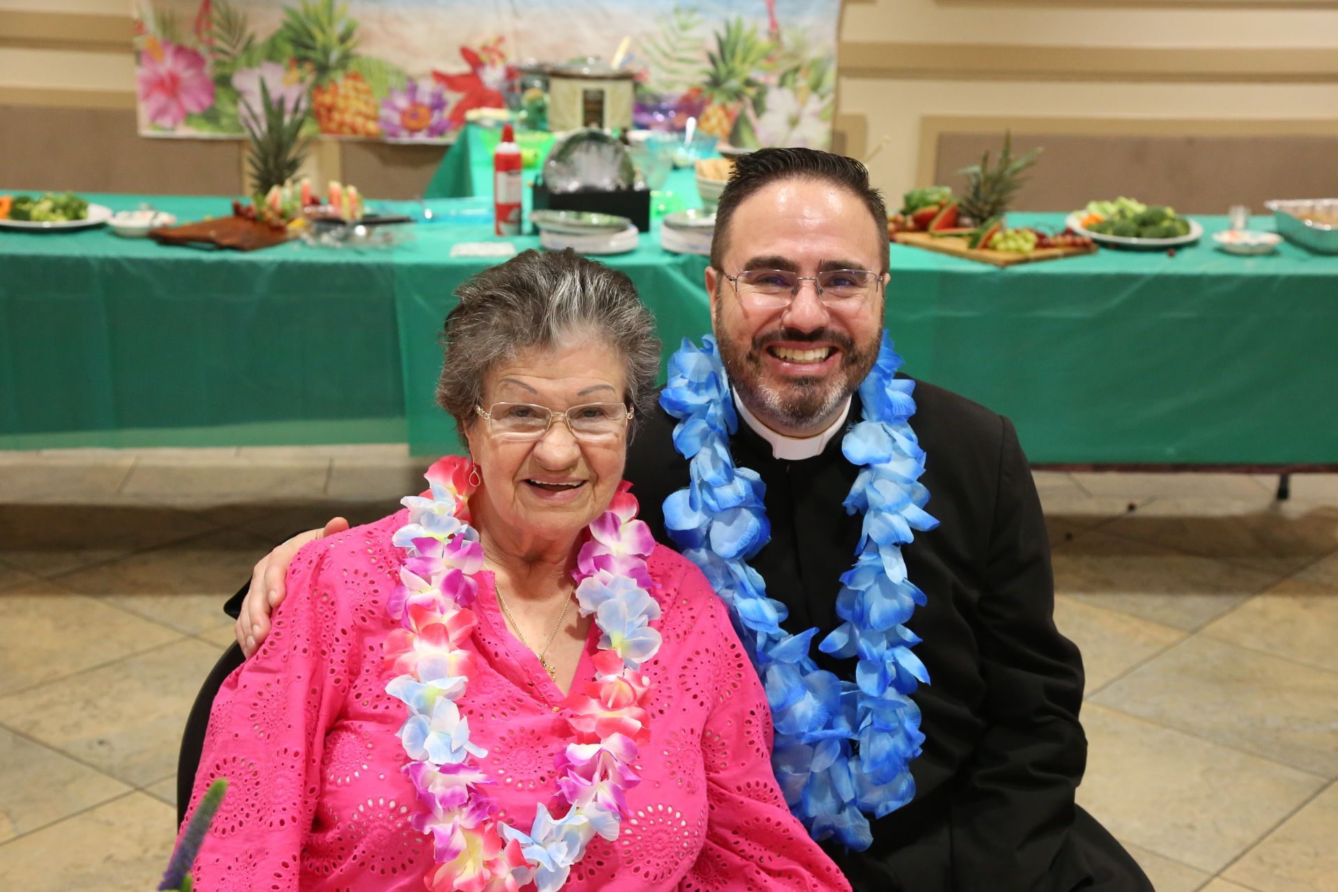 Two smiling people wearing pink and blue leis sit in front of a decorated table at an event.