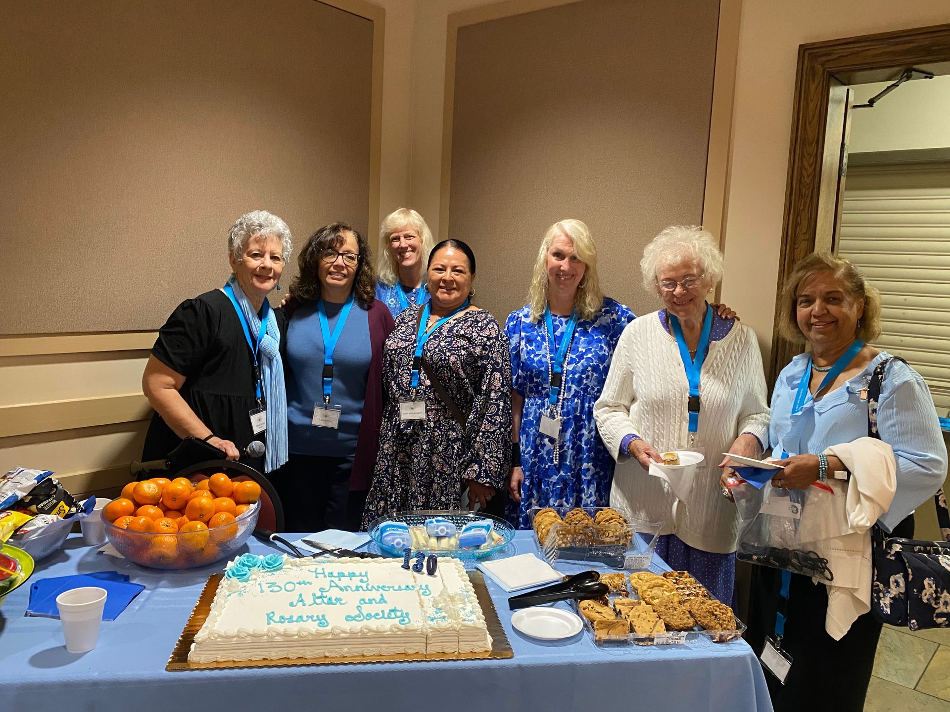 Group of women standing behind a table with cake, pastries, and fruit at an indoor celebration
