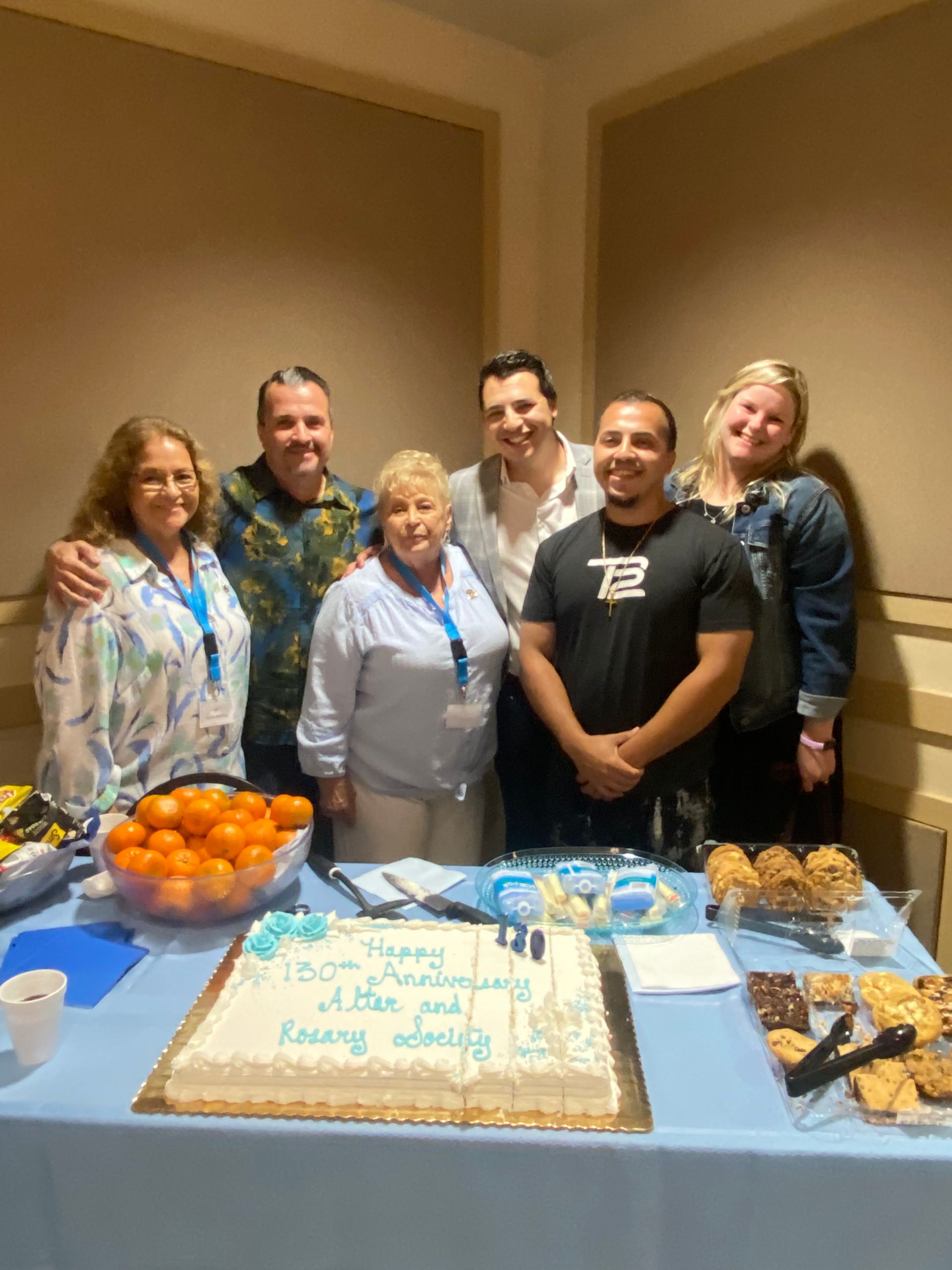 Group posing behind a birthday cake and snacks at a party table indoors.