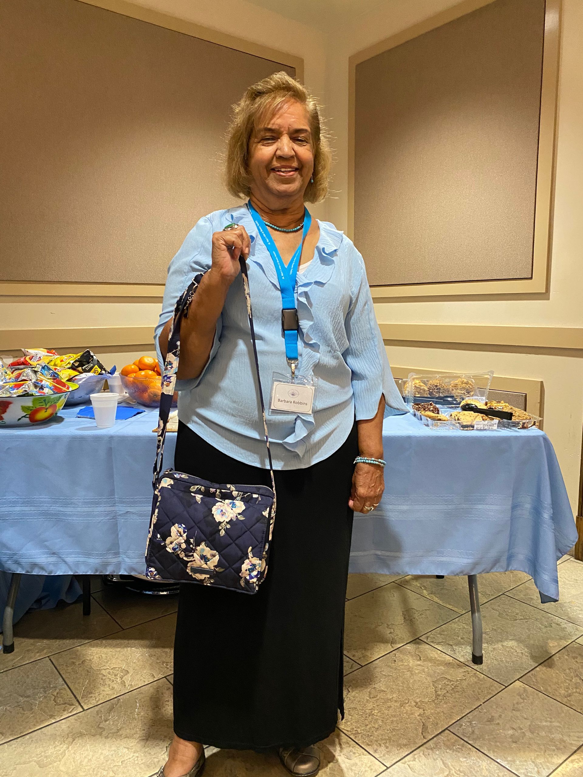 Woman standing indoors beside a table of gifts, wearing a blue blouse, black skirt, and floral shoulder bag.
