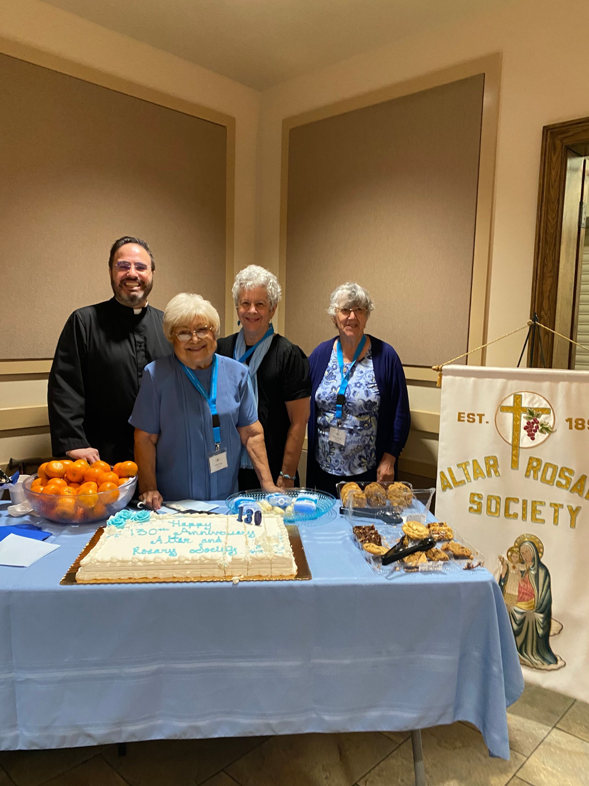 People standing behind a decorated cake table at a community event, with a banner and flowers.