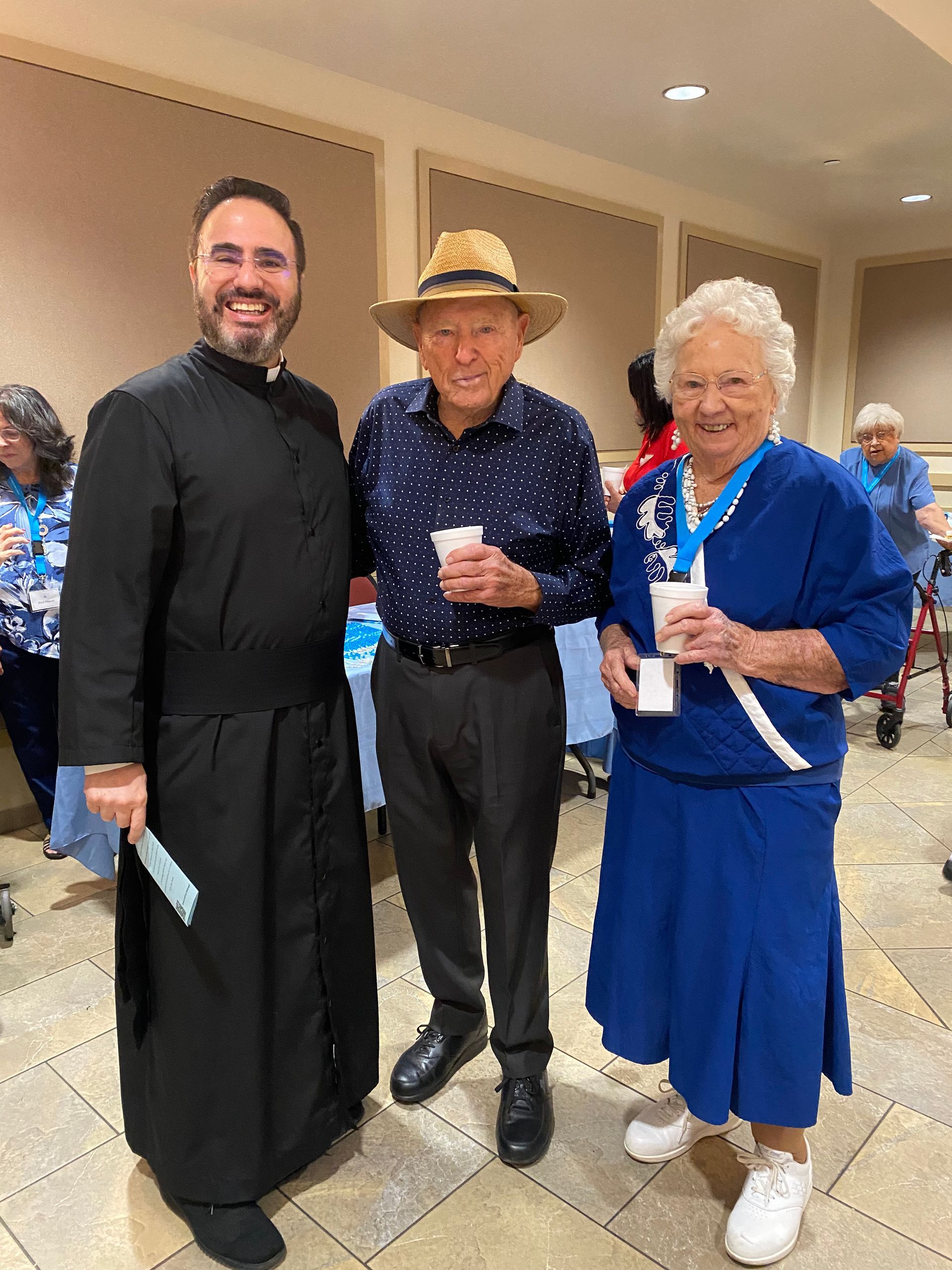 Three people posing indoors at an event, two holding drinks, one in a black robe and two in blue outfits.